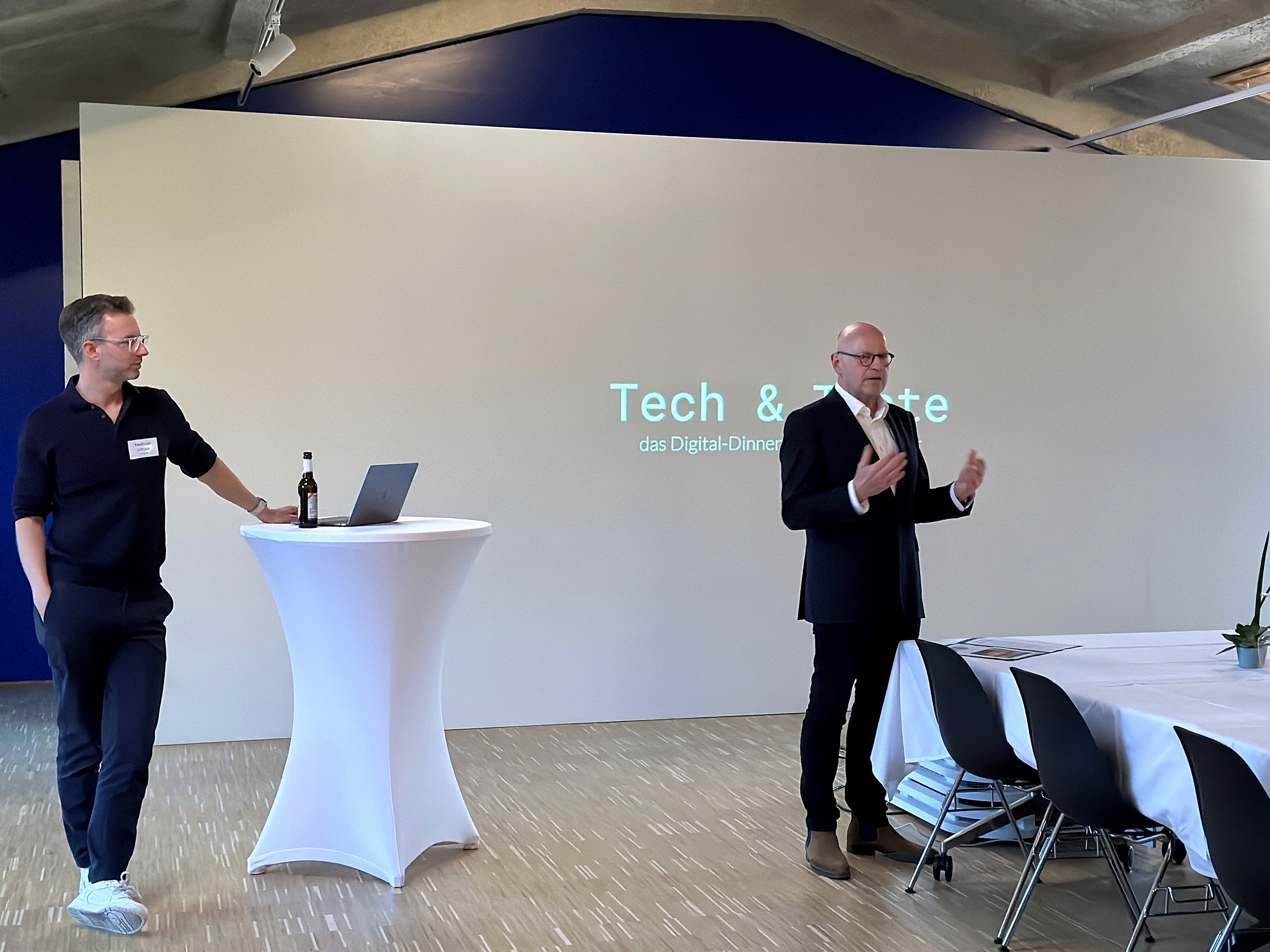 Vortragsszene in einem modernen Raum mit Leinwand, Stehtisch und Stuhlreihen; zwei Personen präsentieren ein digitales oder technologisches Thema.