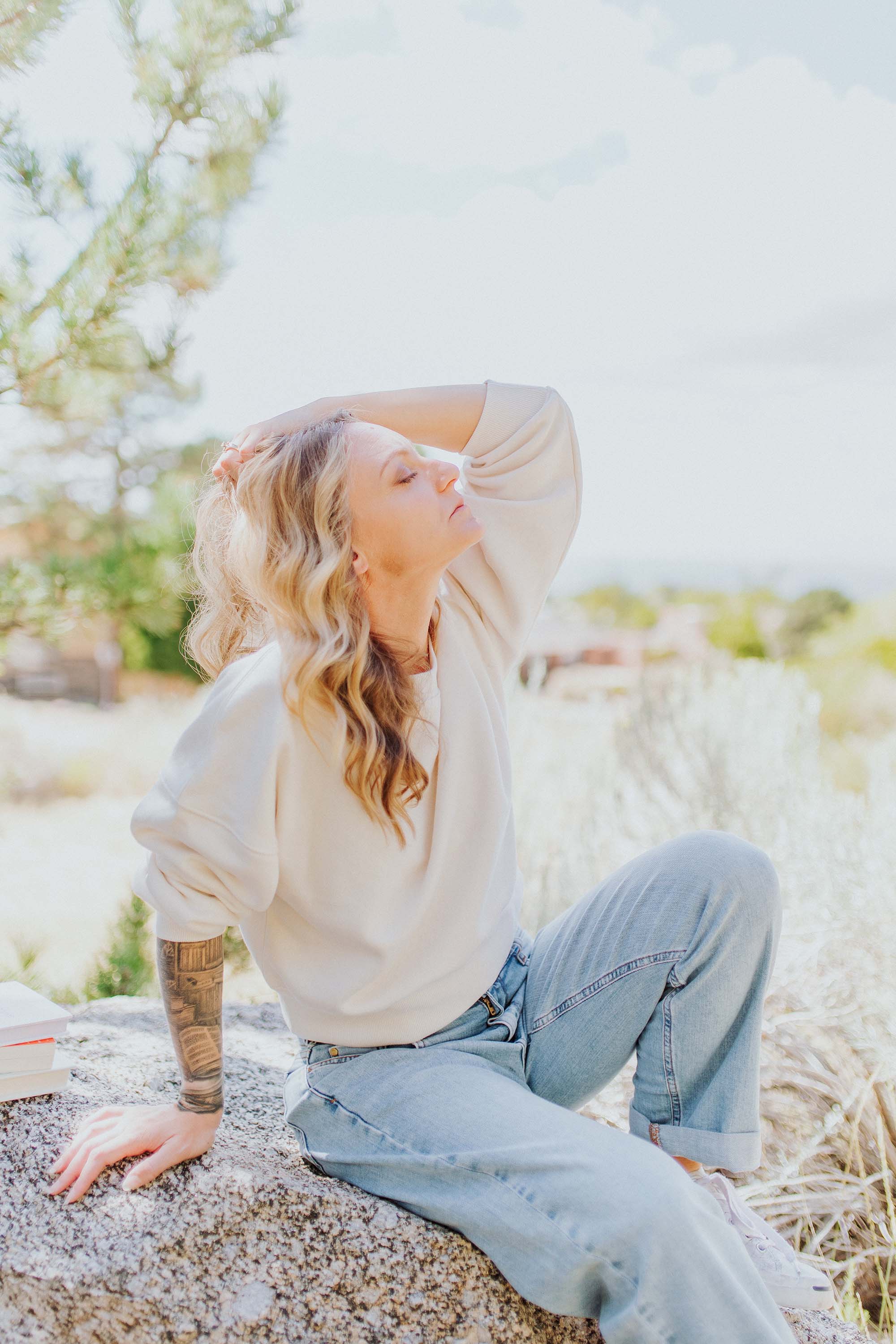 A white woman wearing a white sweatshirt and light blue jeans with wavy strawberry blonde hair is sitting on a rock outside, her face is to the sky and she is leaning back on one tattooed arm