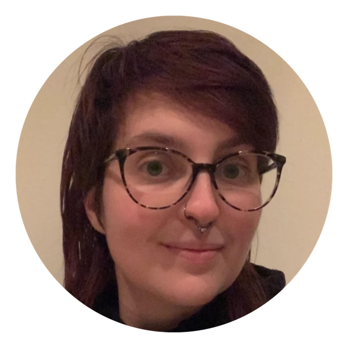 A circular portrait shows a person with shoulder‑length dark hair and glasses in a neutral indoor setting. Soft lighting and plain walls keep the focus on the subject.