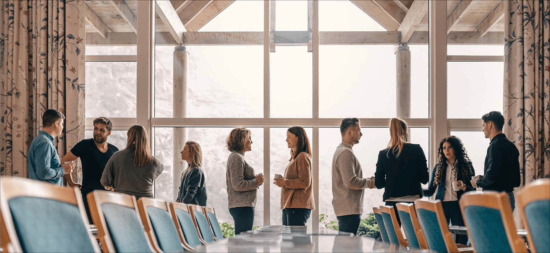 A group of people gathered in a bright room with large windows, engaged in lively conversation, creating a professional networking atmosphere.
