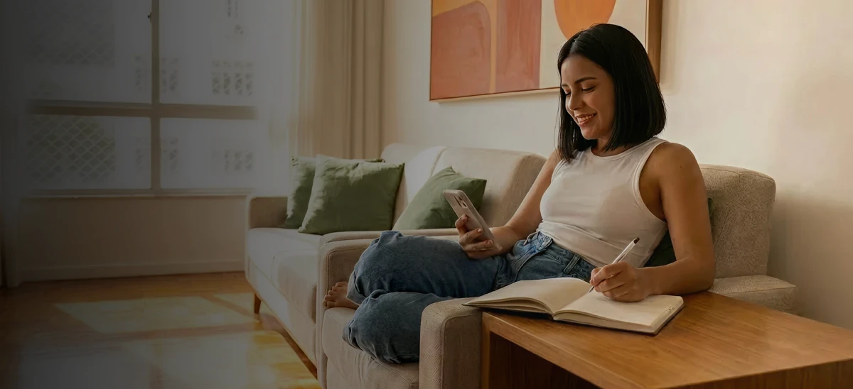 Mulher sentada no sofá escrevendo no caderno e olhando o celular