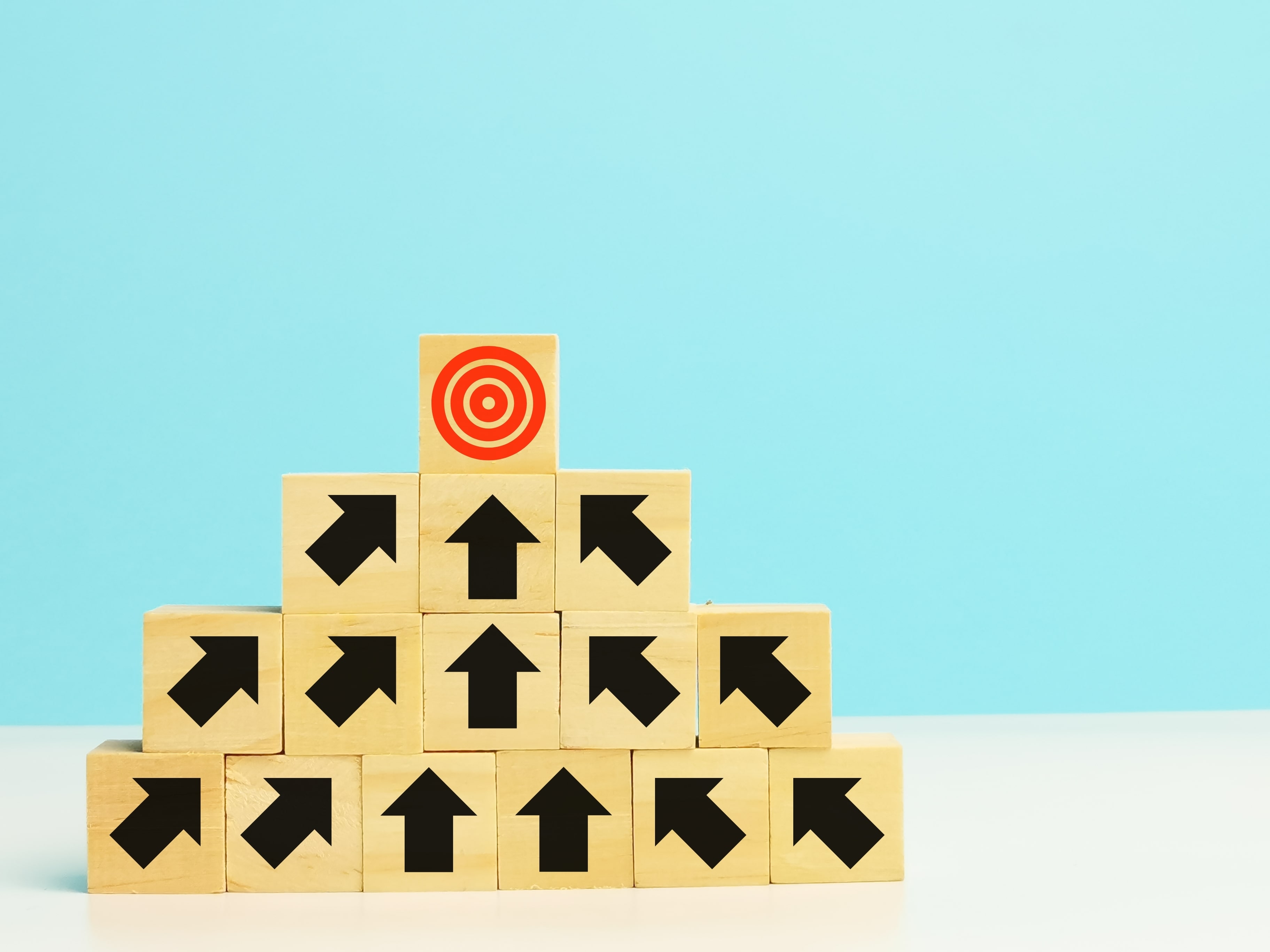 Pyramid of wooden blocks with upward-facing black arrows pointing toward a red target icon on the top block, symbolizing goal setting, strategic planning, and achieving business objectives on a blue background.