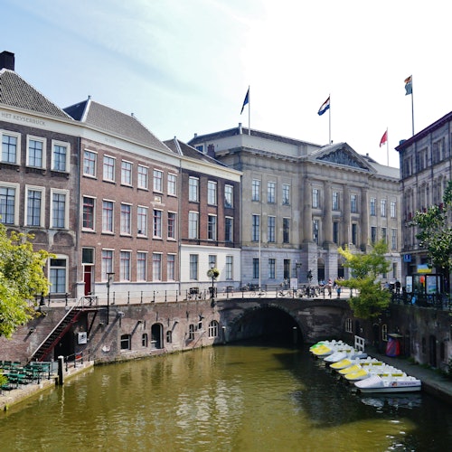 Cyklar parkerade längs en kanal med trampbåtar på vattnet, kantad av historiska byggnader och en valvbro i bakgrunden.