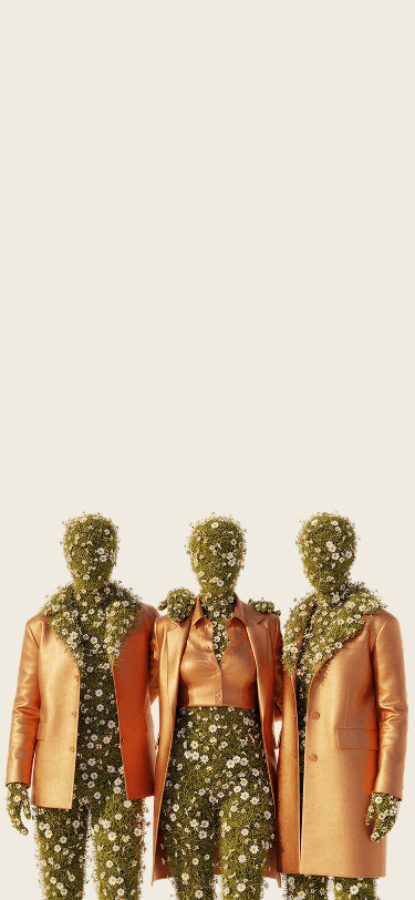A surrealist digital artwork of three humanoid figures standing side-by-side against a cream background. Each figure is composed of green grass and blooming white daisies where skin would be, including their heads and hands. They are dressed in fashionable, metallic bronze-orange suits and a crop-top blouse. The figure in the center has their arms around the shoulders of the other two, and daisy vines spill over the lapels and collars of their jackets.