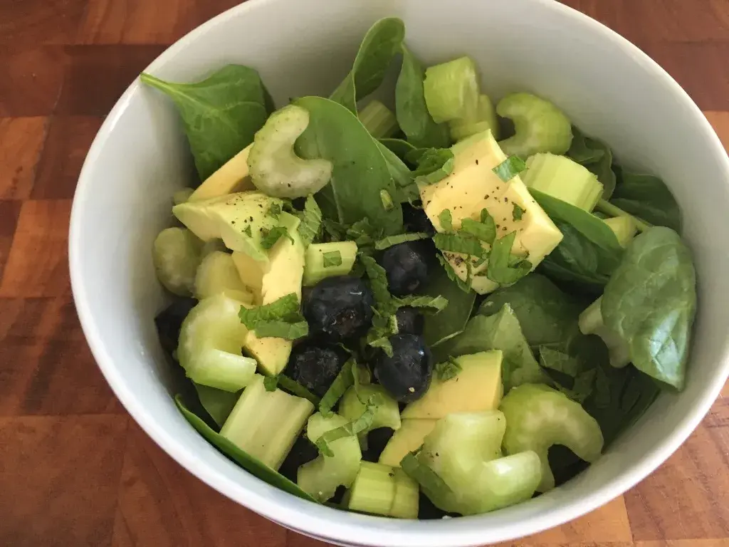 A fresh salad bowl featuring vibrant green spinach leaves, sliced creamy avocado, crunchy celery, and plump blueberries, garnished with a sprinkle of chopped herbs, rests on a wooden surface.