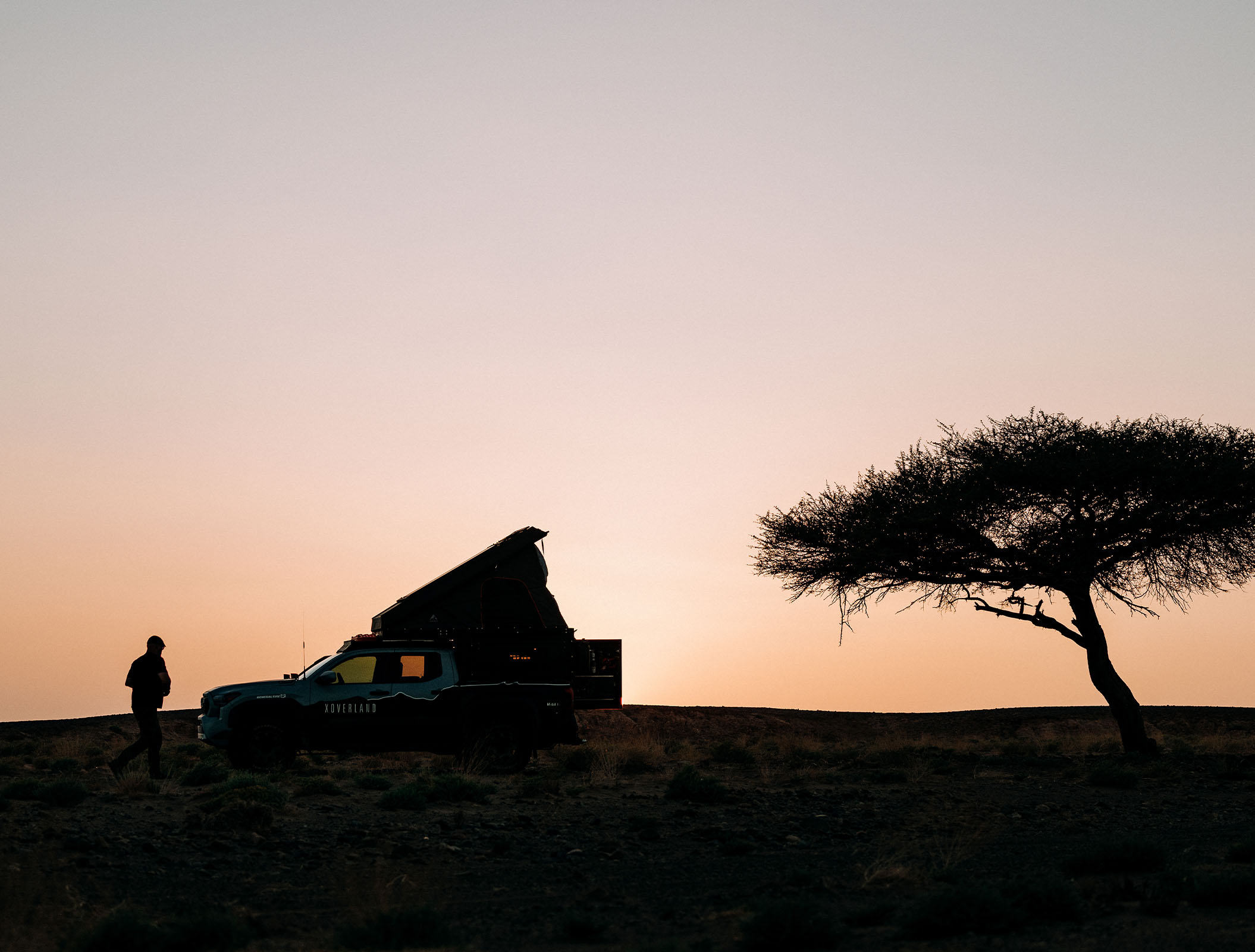 Silhouette of an overland vehicle with a rooftop tent and a person standing beside it on a ridge at sunset, with a lone tree to the right against a clear sky.