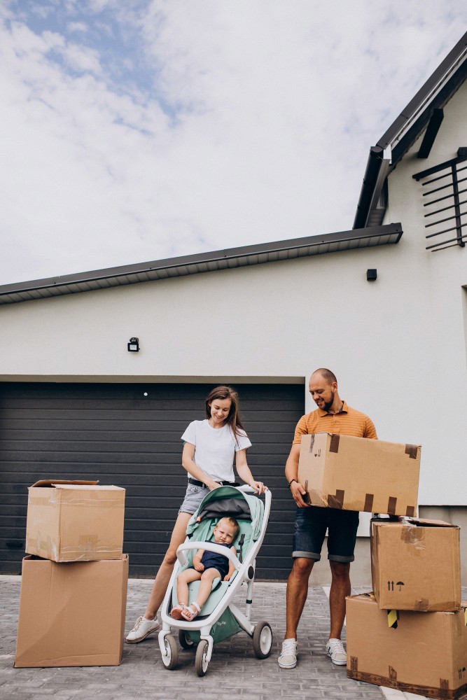 A young family stands outside their new home surrounded by moving boxes, with the mother pushing a stroller and the father carrying a box. The image represents the excitement and fresh start made possible through USDA Rural Development loans from Chris Lewis Home Loans.