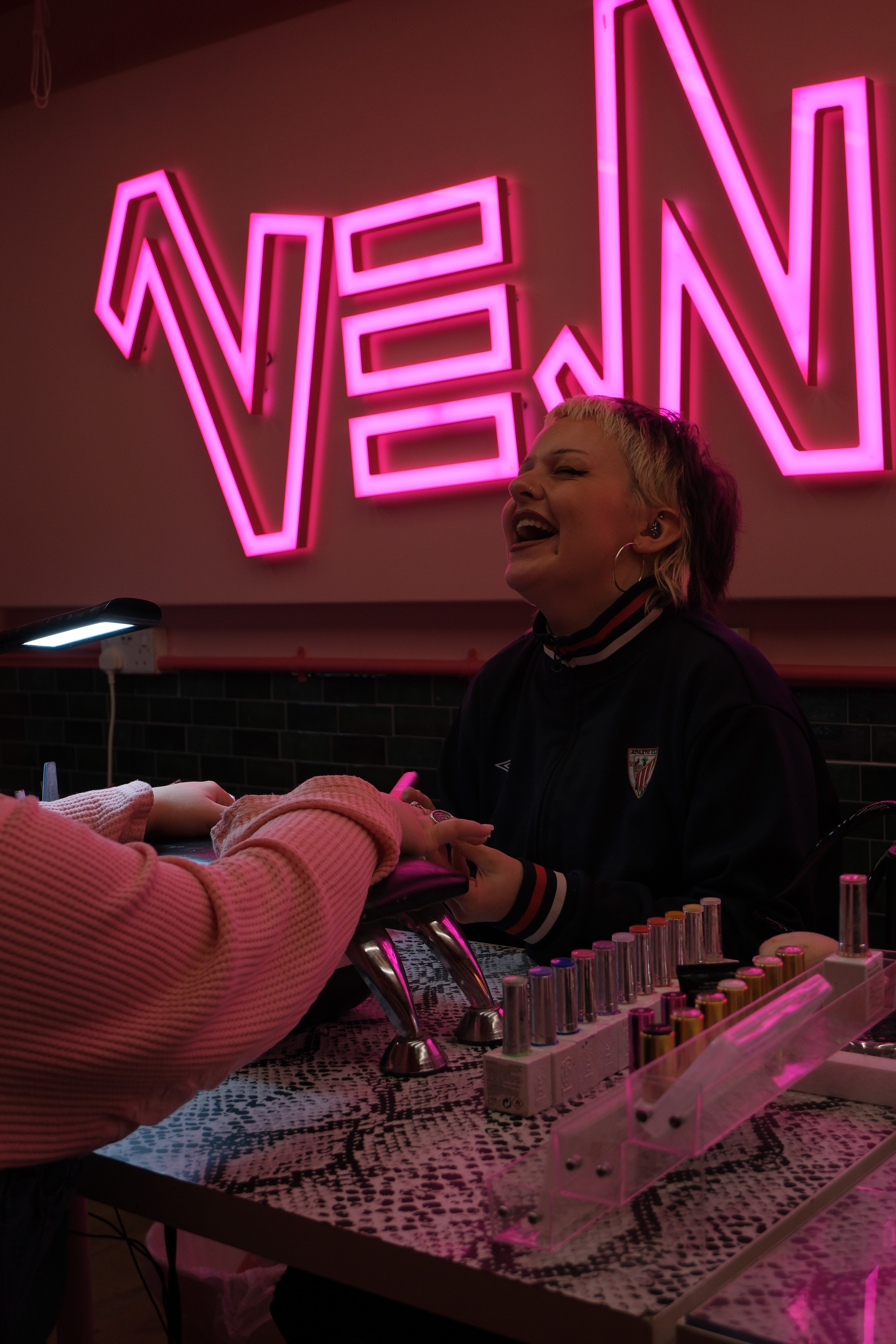 A candid shot of a nail technician with blonde-streaked hair and gold hoop earrings, laughing while performing a manicure. A massive pink neon "VENOM" sign glows behind her.