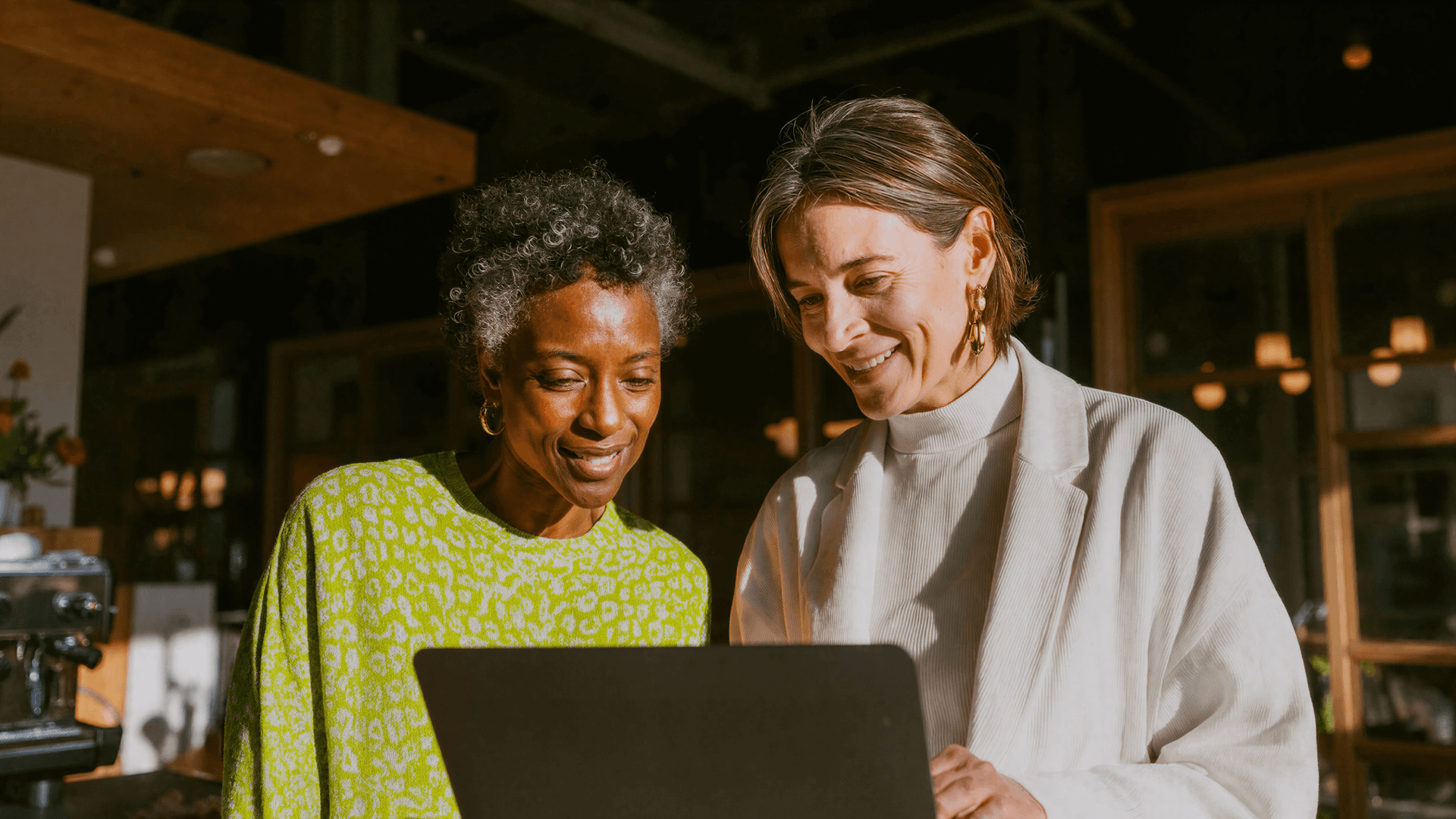 Fotografia em plano médio de duas mulheres em um ambiente de clínica sofisticado, banhado pela luz do sol. À esquerda, uma mulher negra de cabelos curtos e grisalhos sorri ao observar a tela de um laptop segurado pela profissional à direita, que veste um blazer branco elegante. O clima é de total harmonia e clareza. O texto alternativo reforça que, com o suporte da Tivita, o aviso de novos valores vigentes na clínica ocorre de maneira humanizada e organizada, deixando paciente e gestão em plena sintonia.