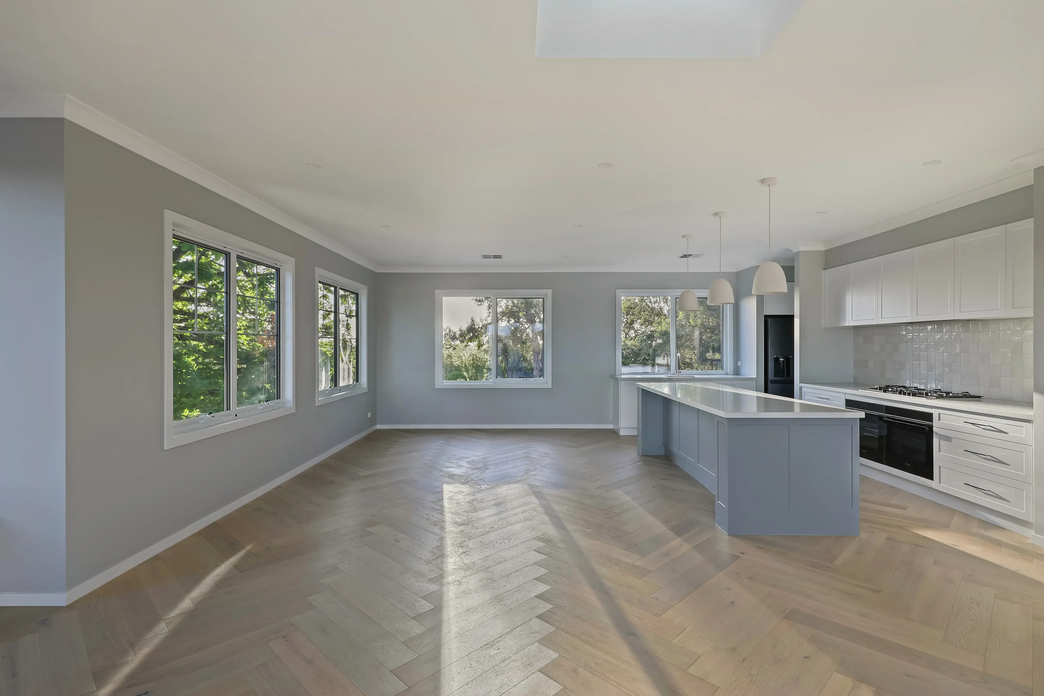 Open-plan living and kitchen with grey walls, white-framed windows to the garden, a grey island with white pendants, gas cooktop, black appliances and oak herringbone floors