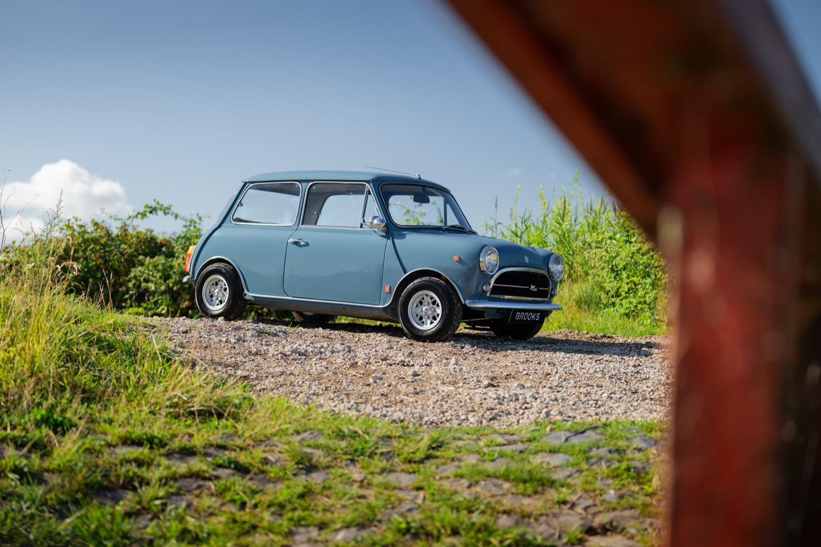 A light blue car parked on a gravel path, surrounded by green grass and blue sky with clouds.