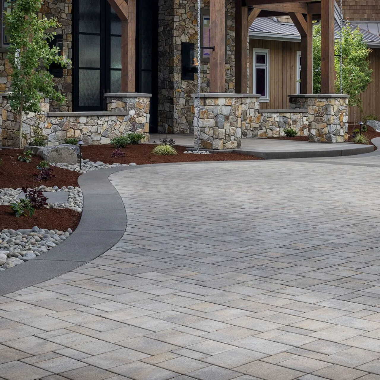 A smooth visual connection Gray paver driveway with a curved dark border beside a stone-clad home.