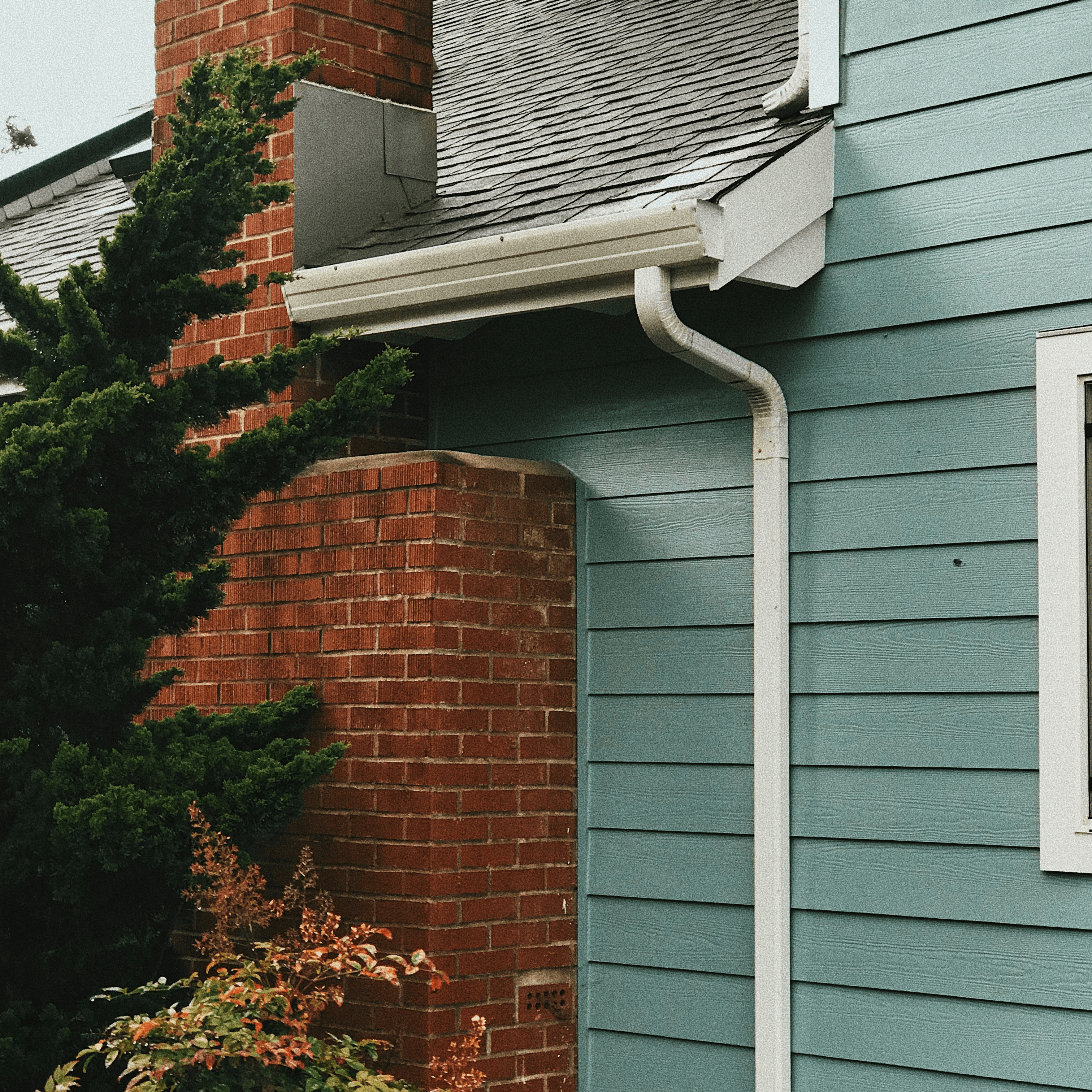 A white gutter downspout on a two story house with light green siding and brick chimney