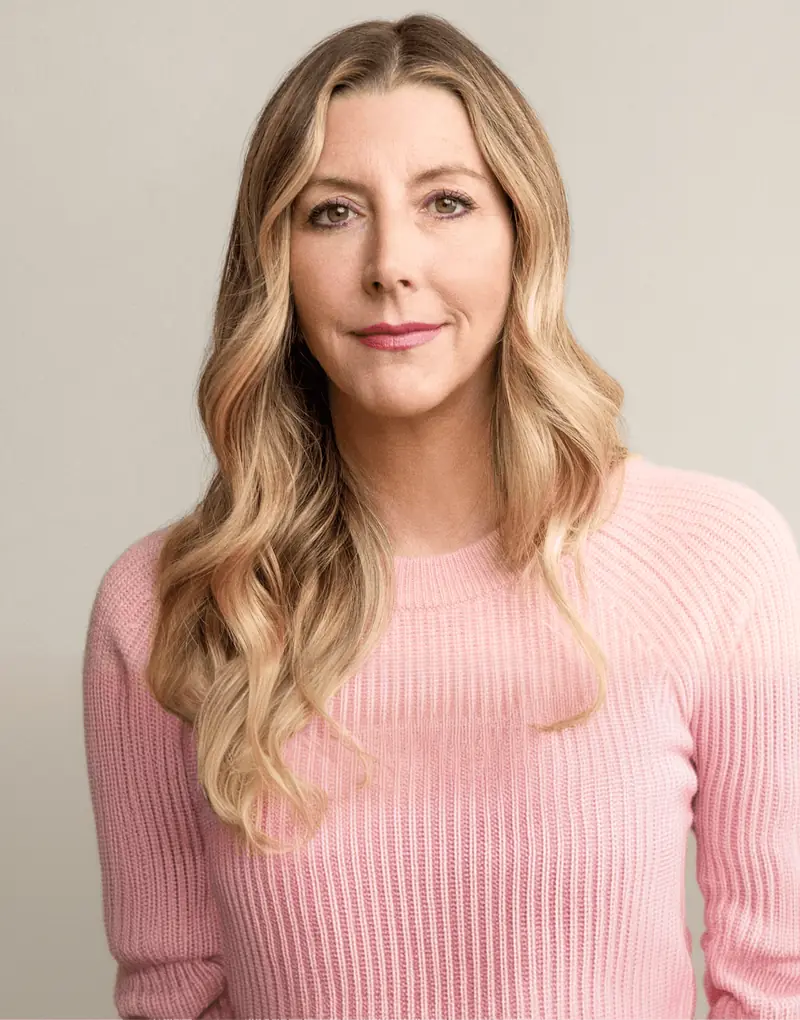 Woman with long blonde hair wearing a light pink sweater, looking directly at the camera with a calm expression against a neutral background.