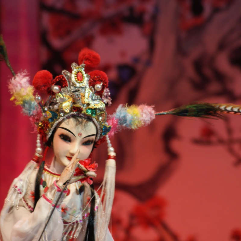 A puppet in a Chinese opera with an ornate head dress, the puppet is being made to delicately smell a flower. The background glows red.