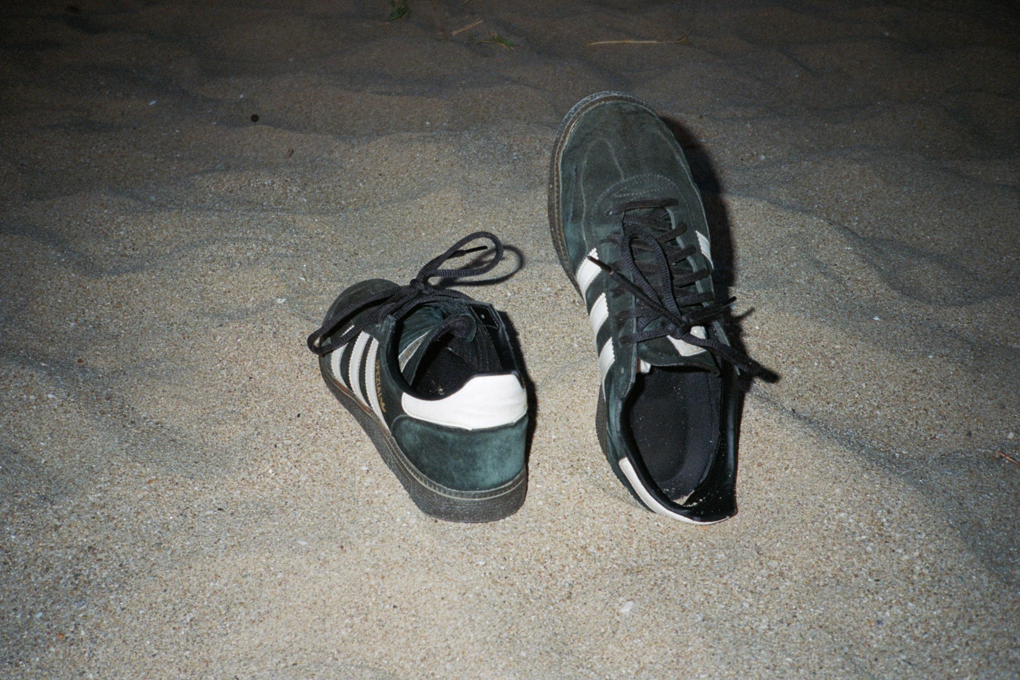 Two green and white sneakers with black laces are placed on a sandy surface, capturing a casual and relaxed beach setting.