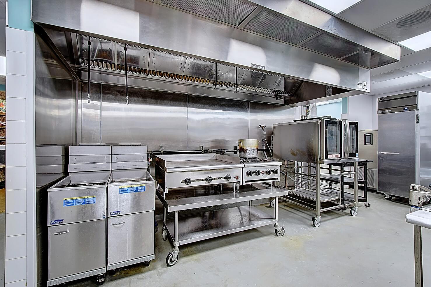 Modern commercial kitchen with stainless steel equipment, including ovens, sinks, and prep tables. Brightly lit space.