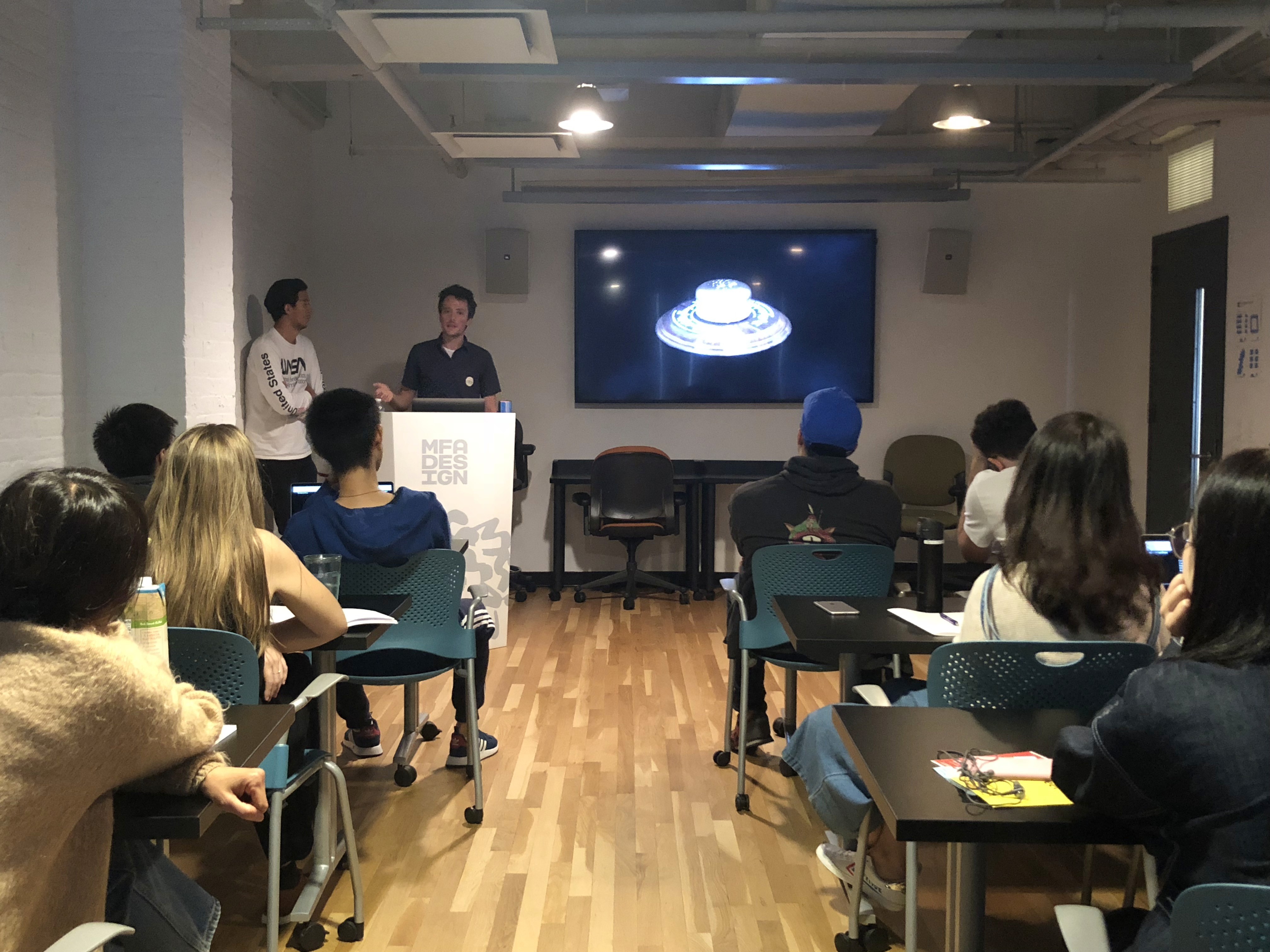Students in a classroom that are watching a white disk shape object on a monitor.