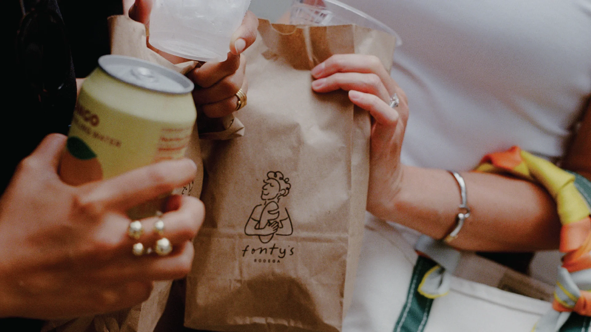 Three people hold brown paper bags with a logo, alongside canned drinks and a clear plastic cup, suggesting a casual takeout meal scene.
