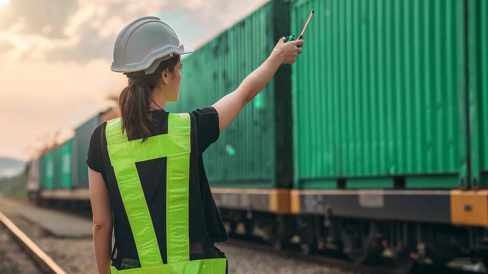 Жінка у касці та жилеті координує відправку зелених контейнерів на залізничному маршруті для доставки вантажів.