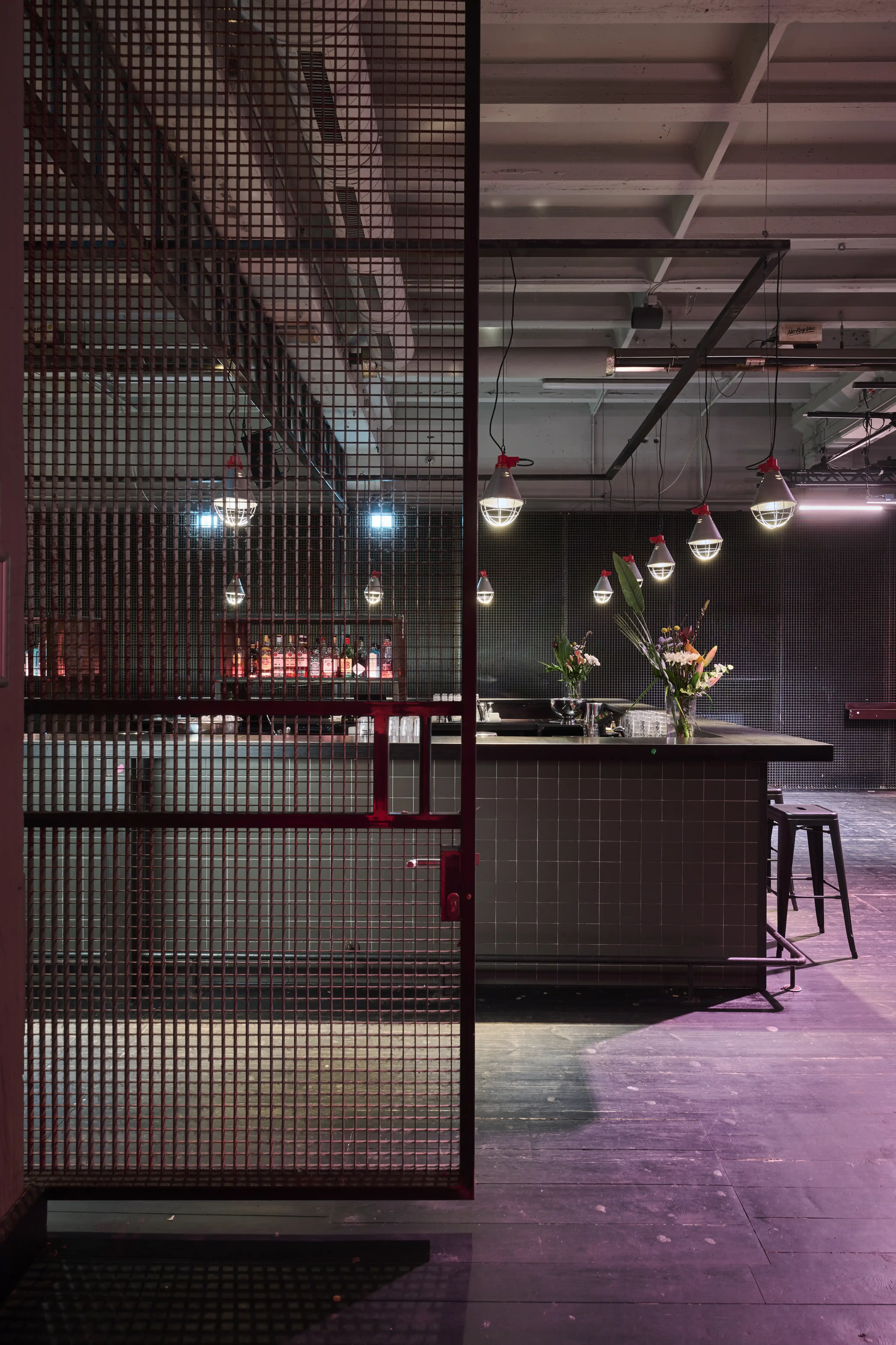 Bar at TRESOR GLOBUS behind a red-framed metal mesh partition, with grey tiled counter, flower vases, glass pendant lights and a white beamed industrial ceiling.