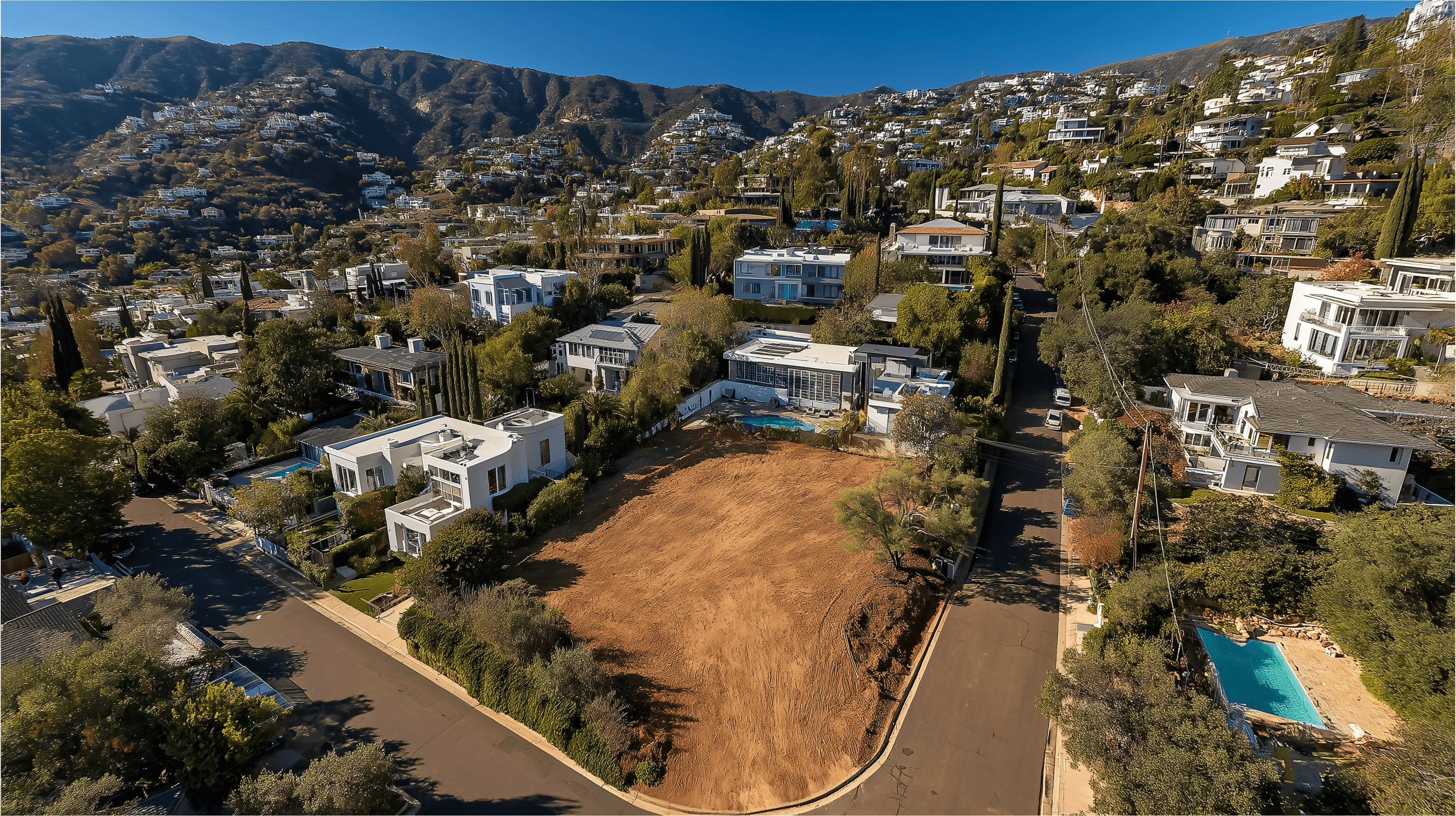 Aerial perspective of a wealthy hillside neighborhood showcasing a large, empty corner dirt lot ready for development, surrounded by modern luxury homes and mature trees.
