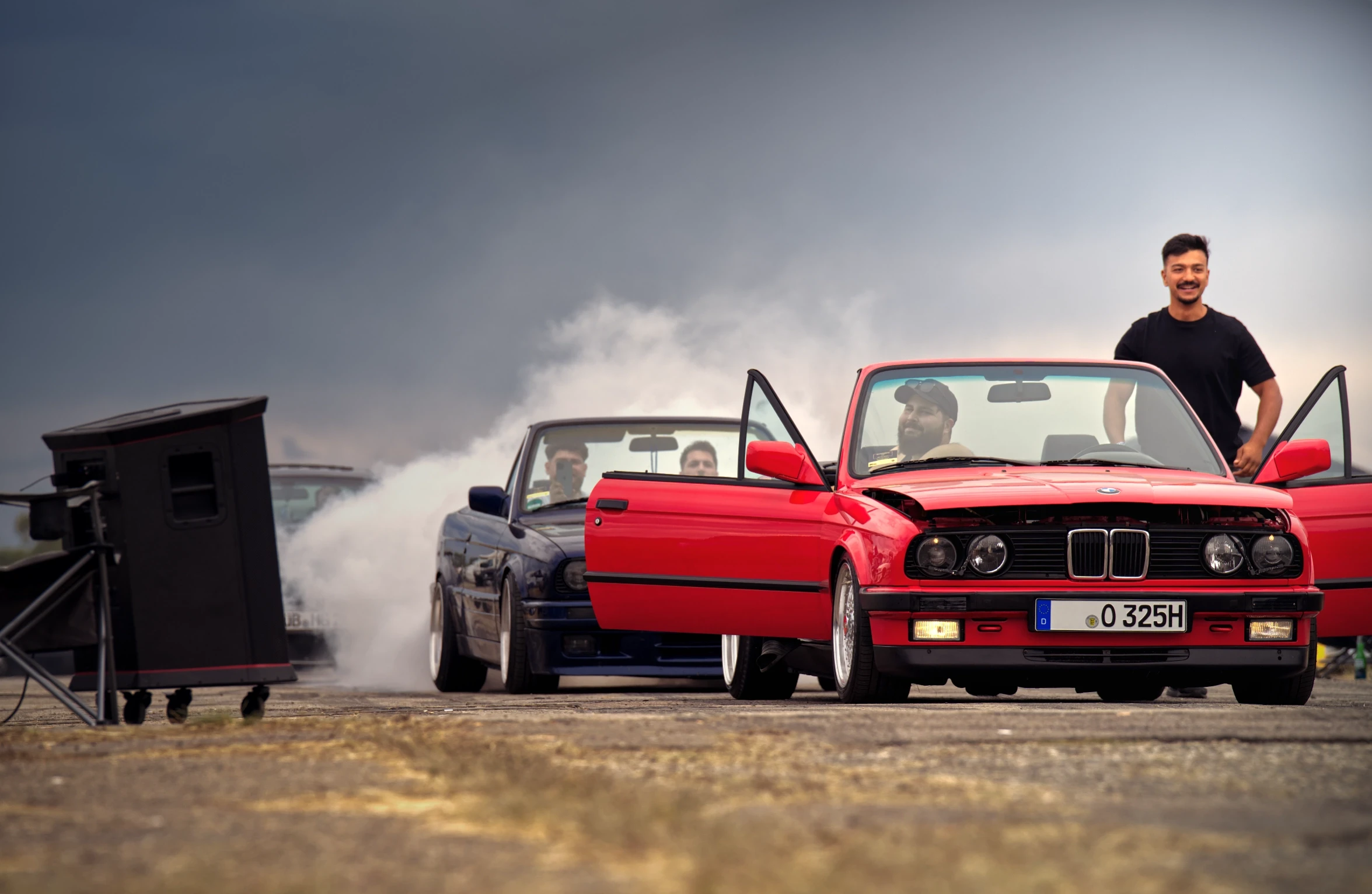 Roter BMW E30 beim Burnout auf dem BMW Syndikat Asphaltfieber 2025 vor dramatischem Gewitterhimmel und aufsteigendem Reifenrauch.