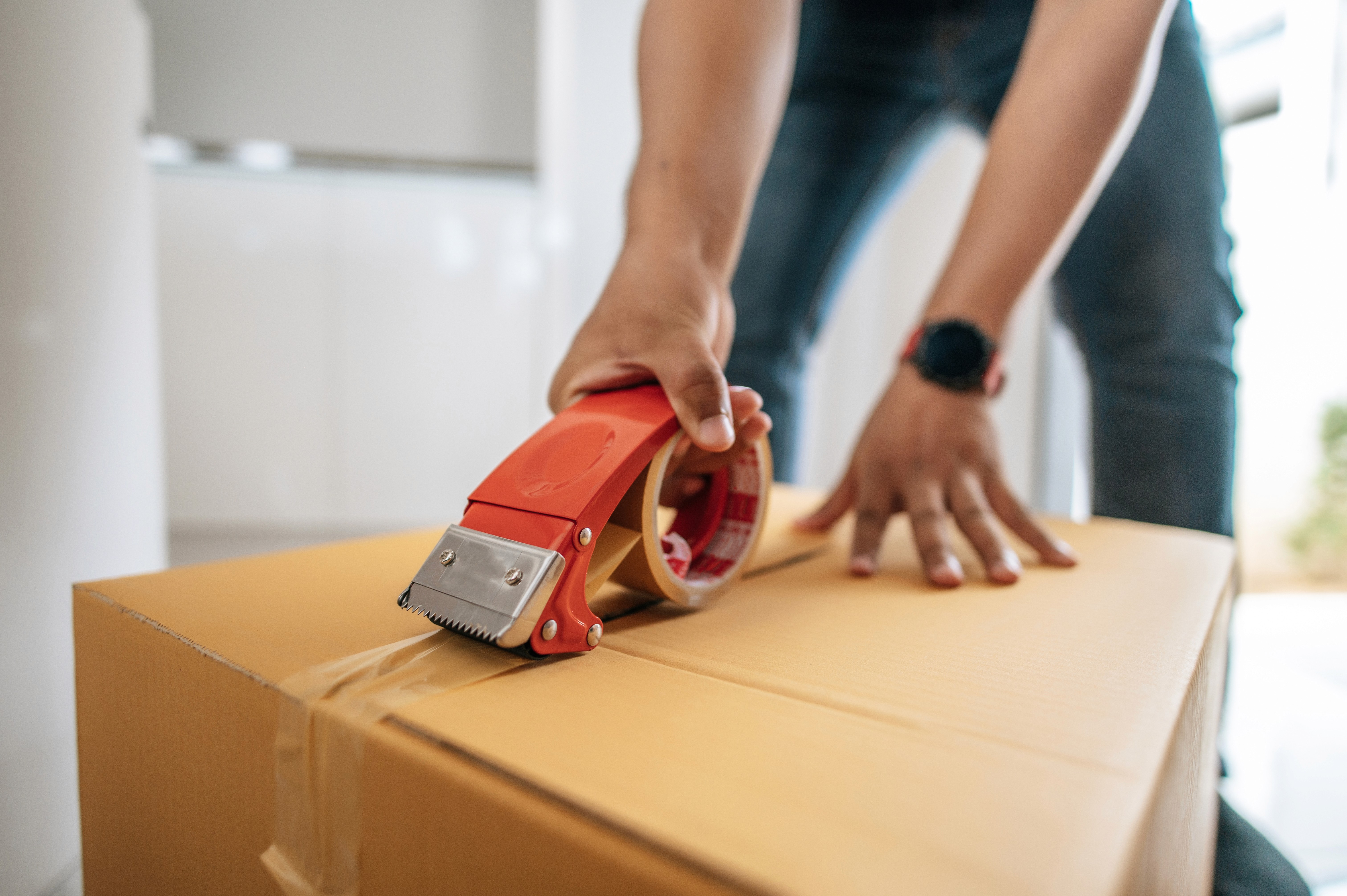 A person uses a red tape dispenser to seal a cardboard box. The scene conveys a sense of moving or packing, with focus on hands and clear tape.