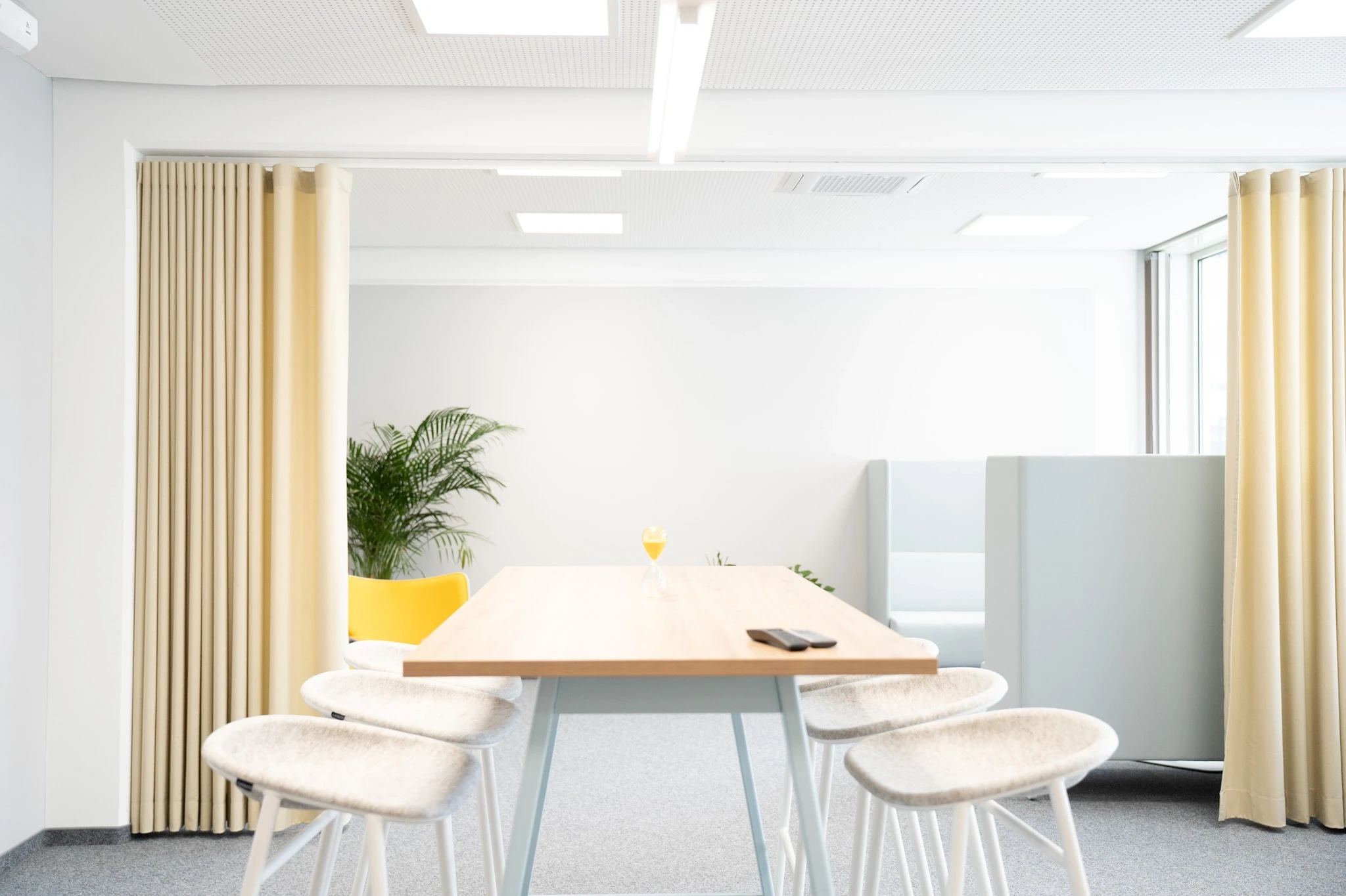 Meeting nook at NFON with a wooden table on pale blue legs, four white furry stools, a yellow armchair, a palm plant and pale yellow curtains framing the space.