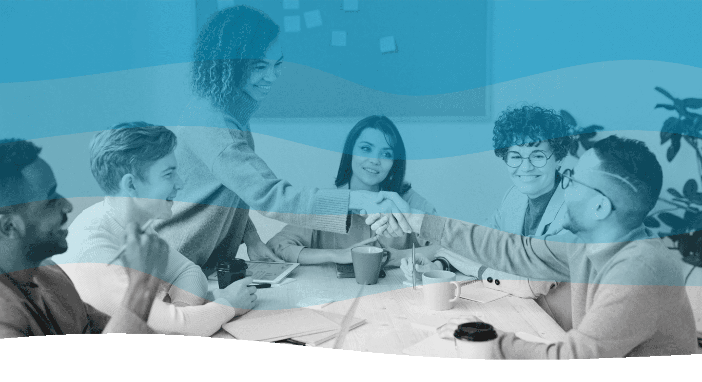 A diverse group of professionals gathers around a conference table, engaged in a meeting, as two individuals shake hands, indicating collaboration and teamwork in a modern office setting.