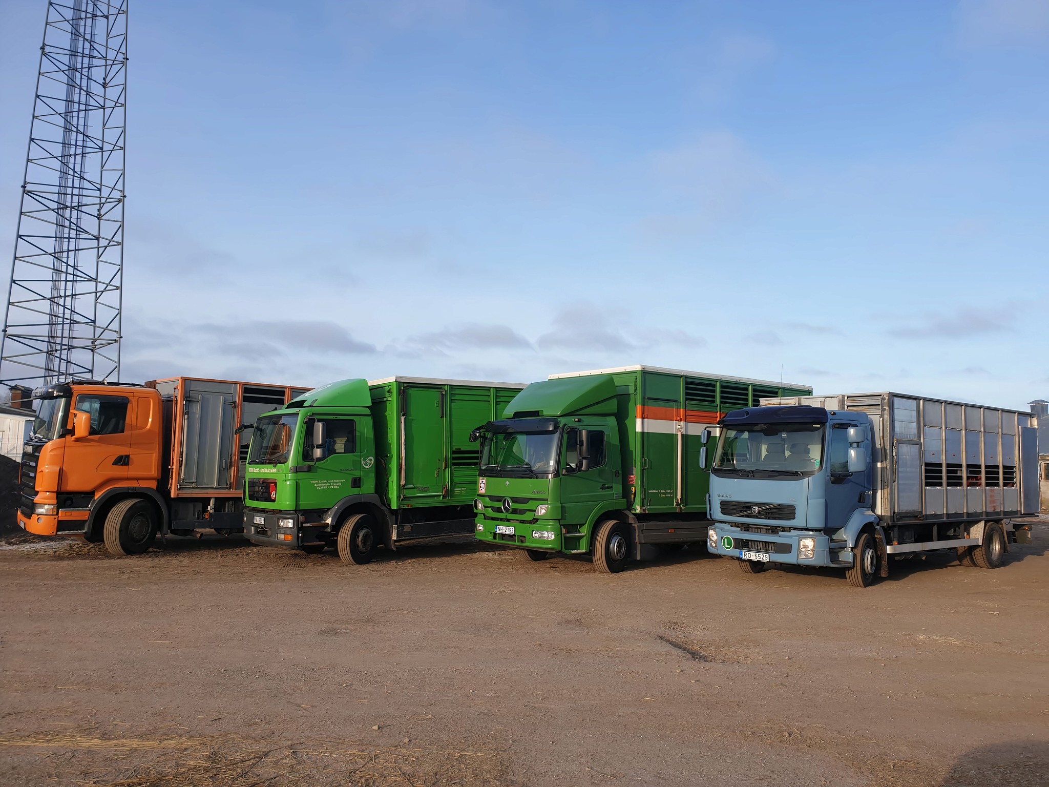 A line-up of four parked trucks in an open lot under a clear blue sky, featuring a mix of orange, green, and blue colors, with a communication tower in the background, highlighting logistics and transportation efficiency.