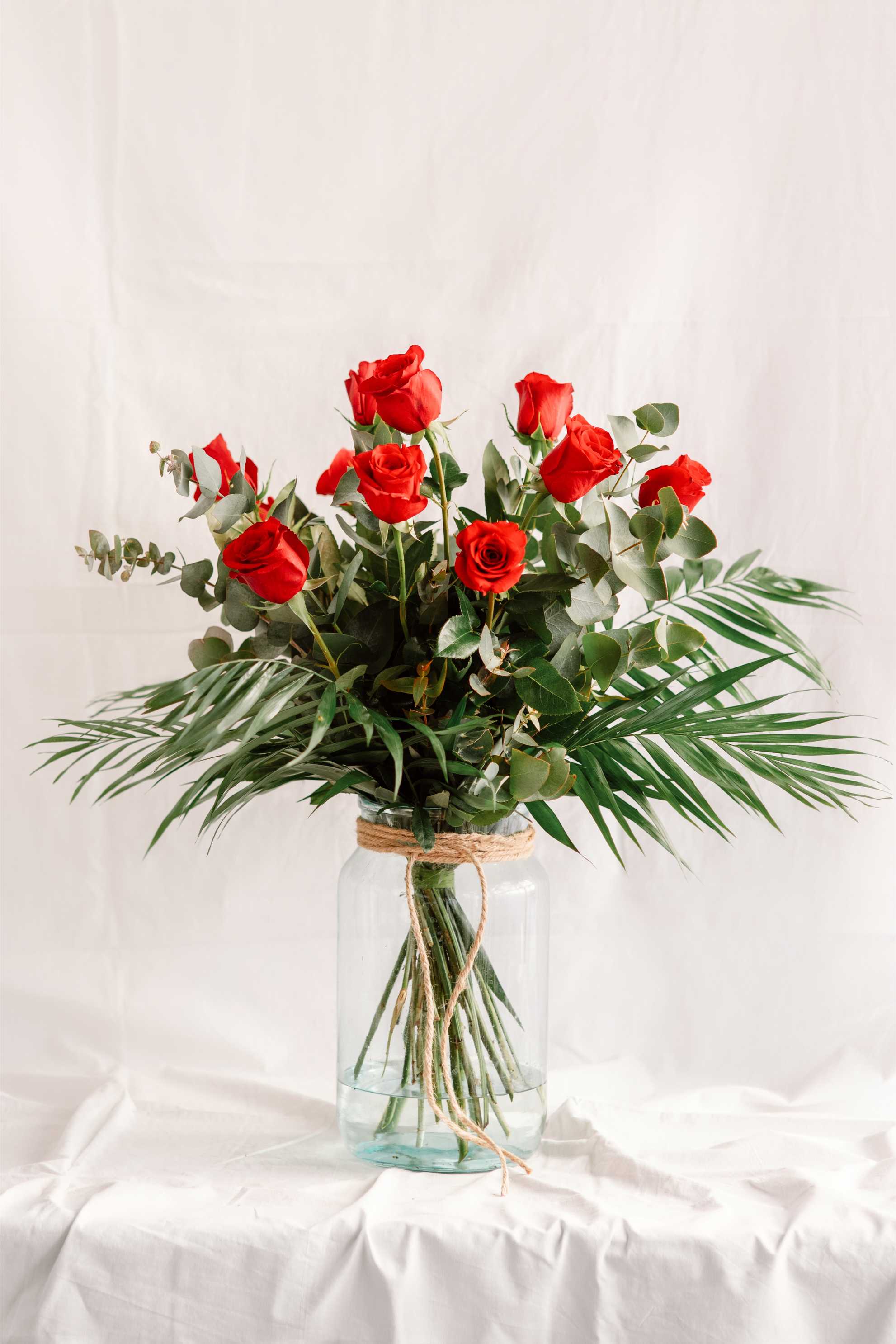 Ram de roses vermelles amb fulles d’eucaliptus i palmera, presentat en un gerro de vidre amb corda natural sobre fons blanc.
