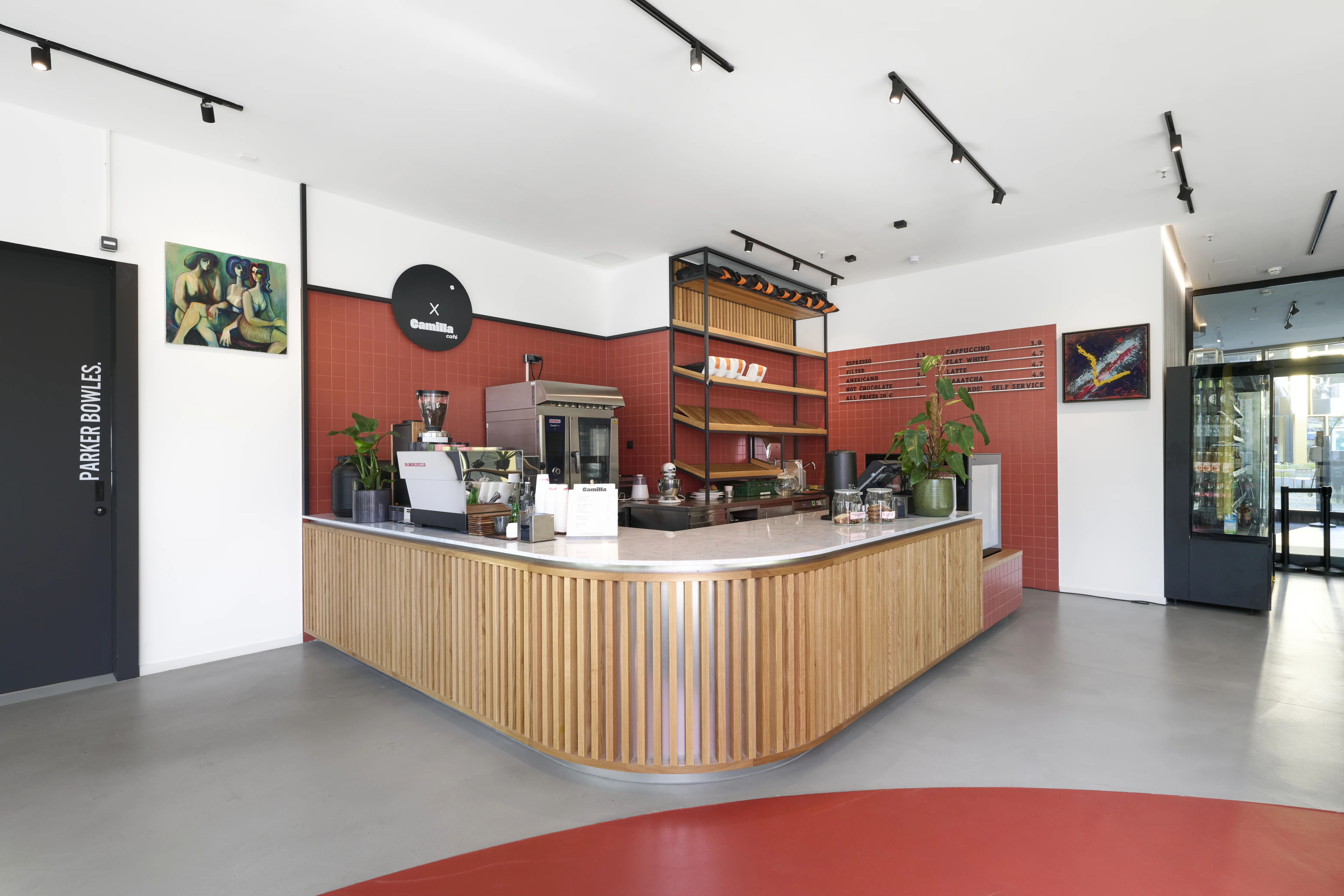 Back counter at CAFÉ CAMILLA with a built-in oven and wooden shelves holding fresh bread, set into a terracotta-red tiled wall.