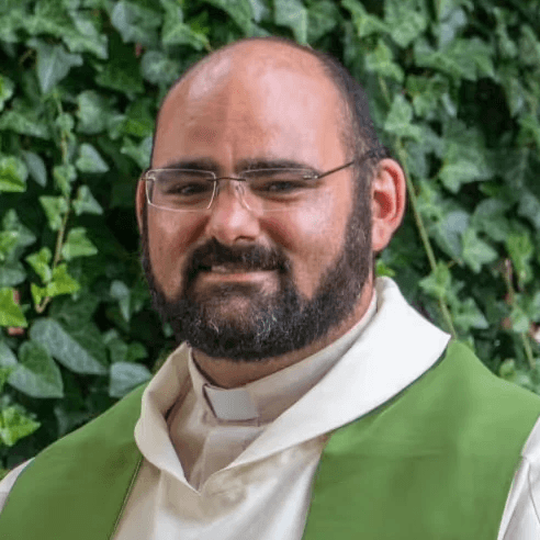 Pfarrer Ludwig Westermeier steht mit Priestergewand vor einer Hecke und lächelt