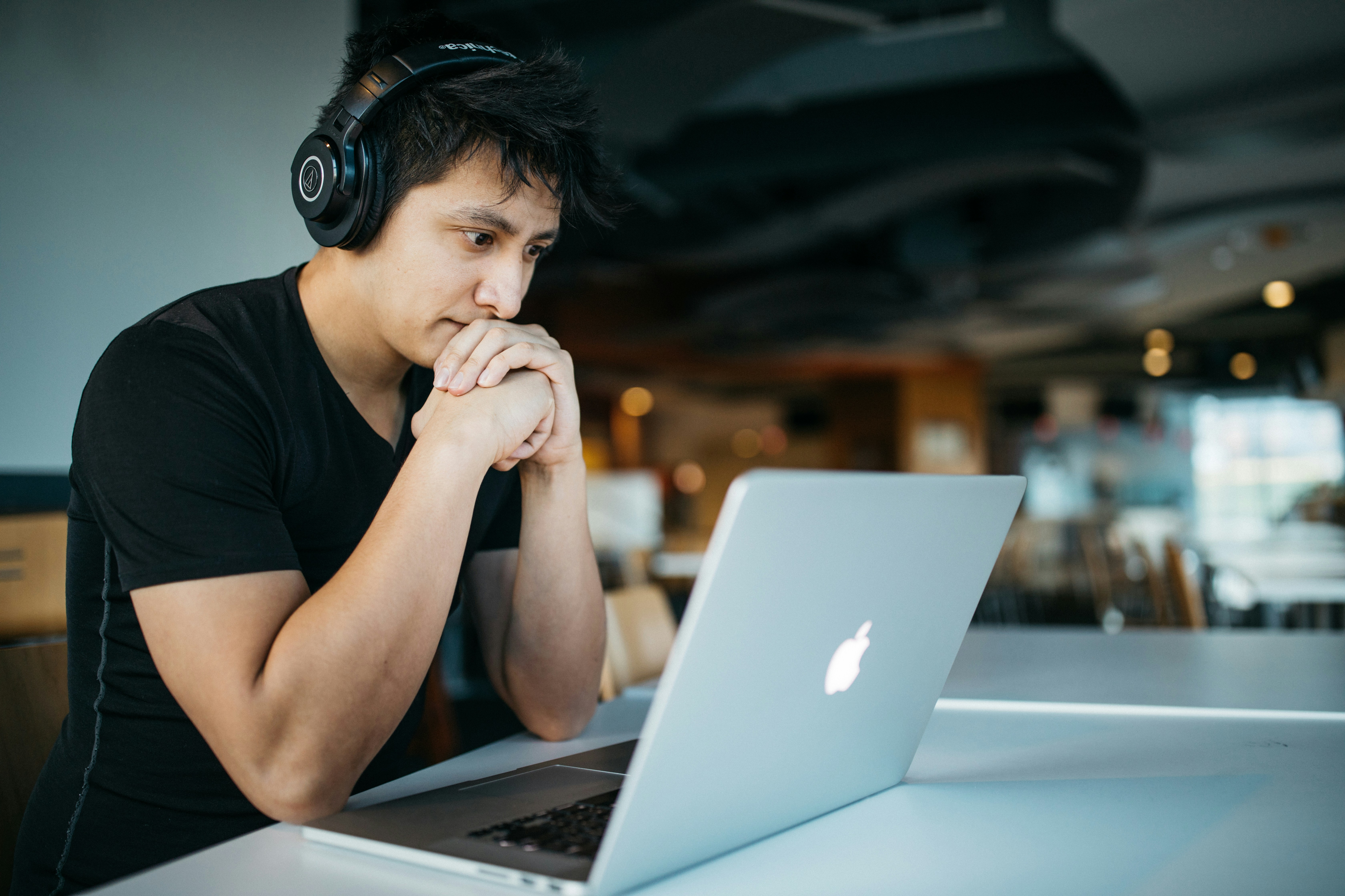 A young man wearing over-the-ear headphones sits contemplating a Macbook, which is on a table in front of him. 