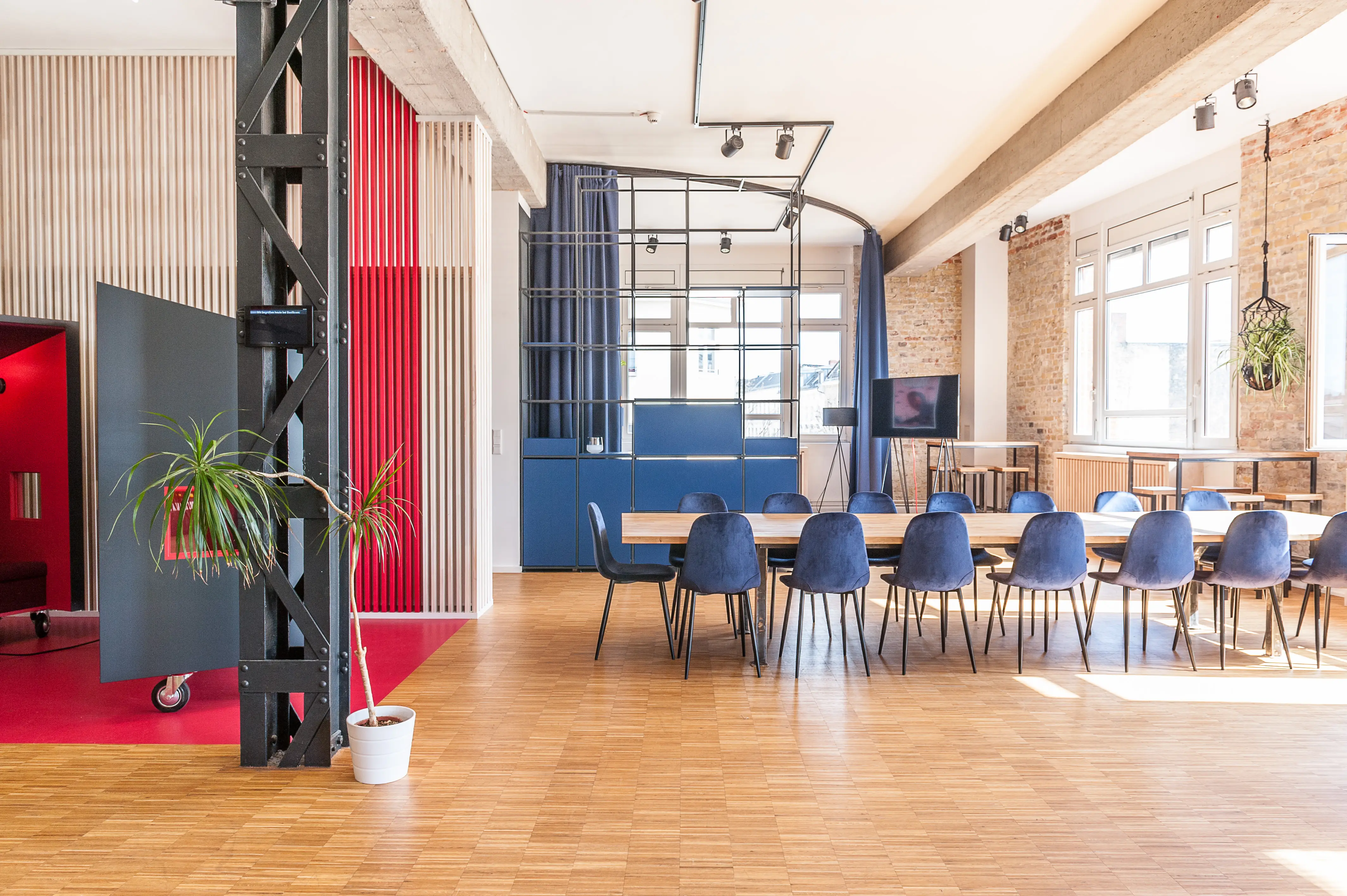 Open loft office at BASILICOM with a long wooden table and petrol-blue chairs, black riveted steel column, red phone-booth boxes, wooden slat walls, exposed brick and large industrial windows.