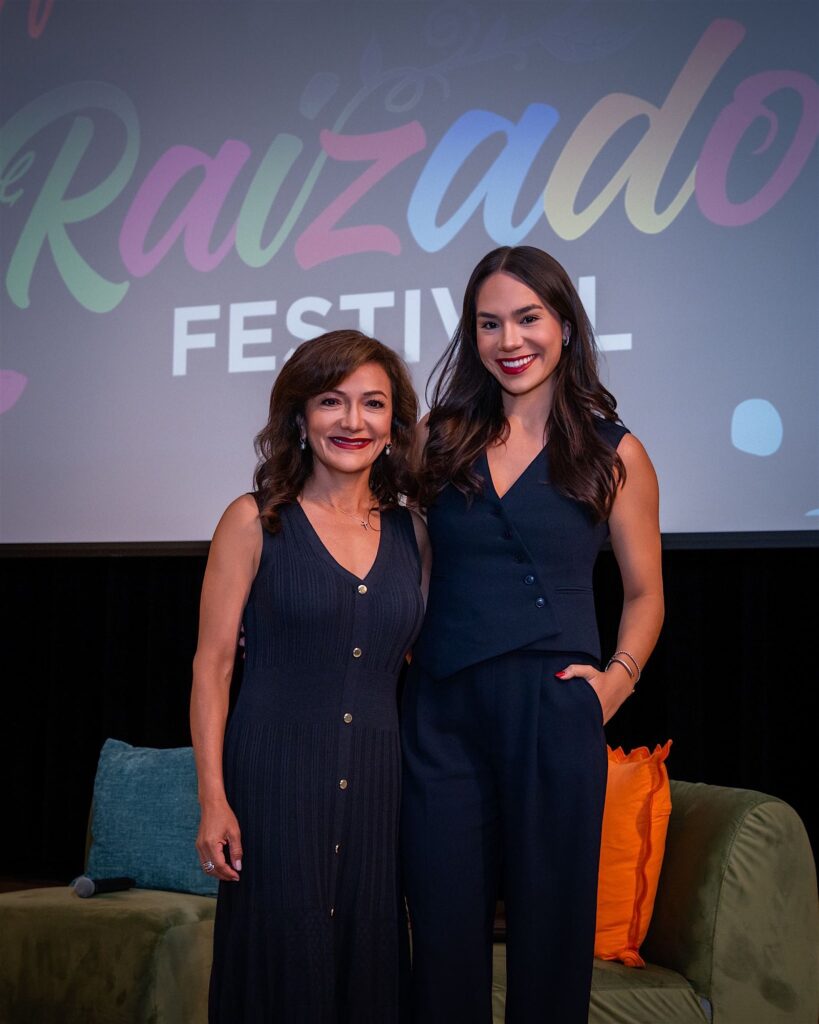 Two guests in navy outfits posing in front of a screen displaying Raizado Festival branding