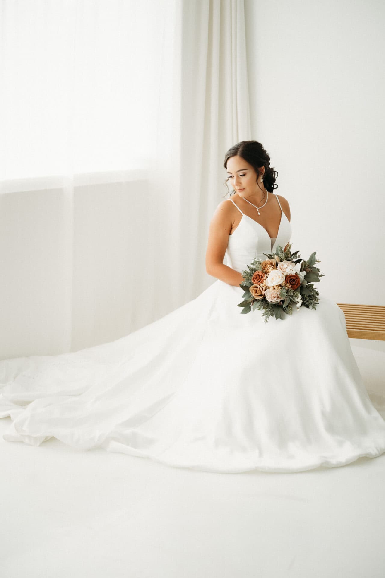 A bride sits by a window in a flowing white gown, holding a bouquet, at Revelator Studio, a natural light photography studio in Shreveport.