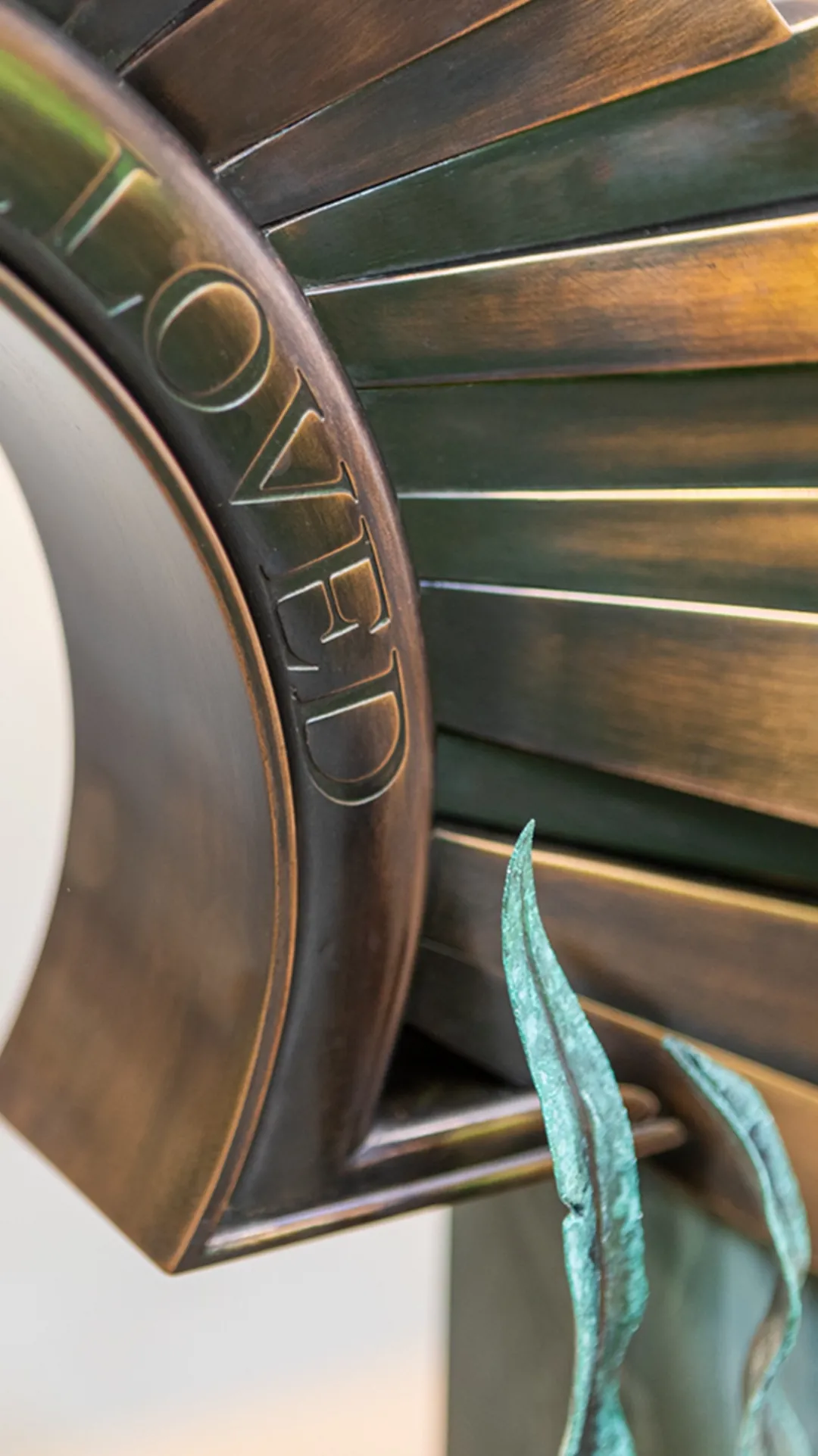 A close-up detail of a bespoke polished bronze headstone sculpture engraved with text, featuring green patinated bronze leaf elements. A tailored memorial objet d’art piece by Shaun Leane.