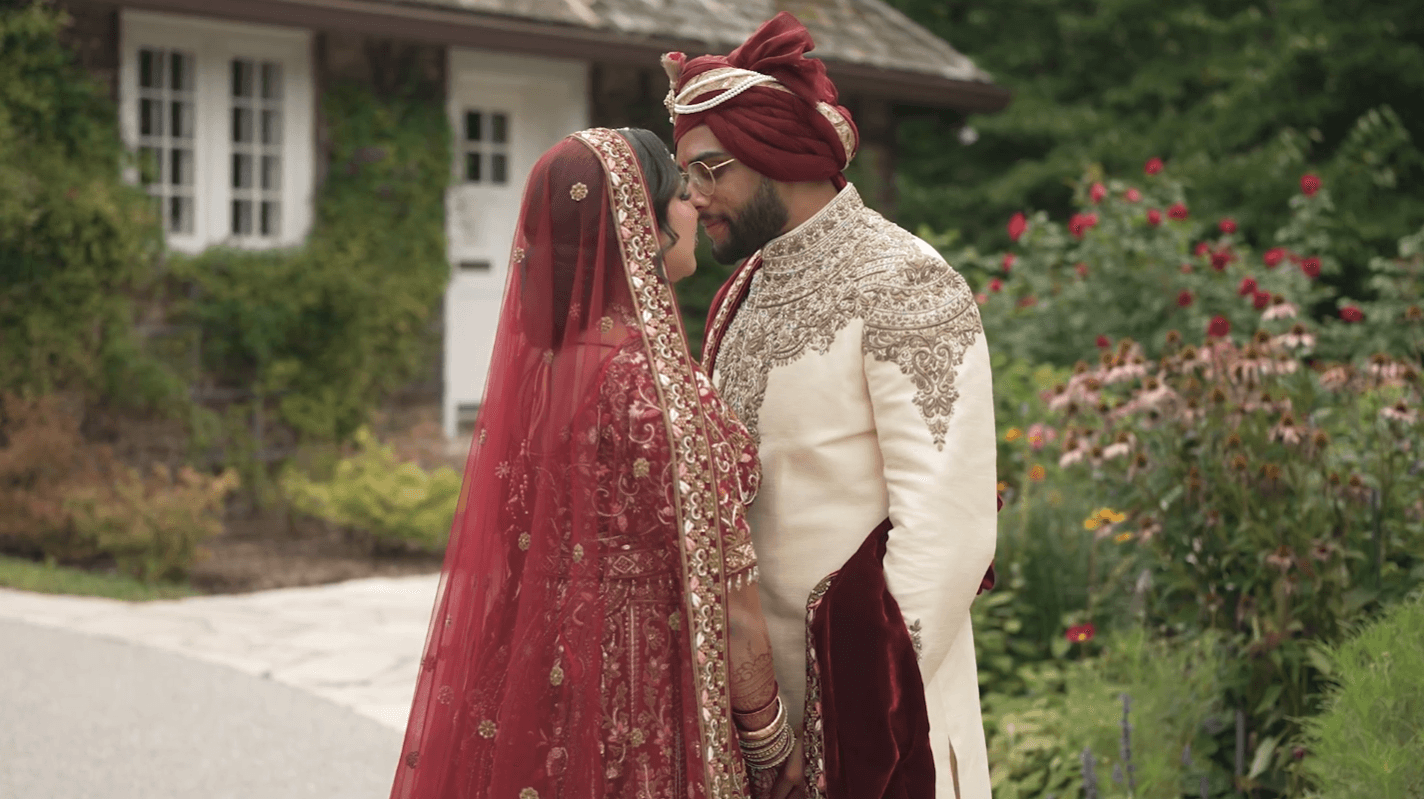 Man in cream and burgundy attire and woman in matching burgundy attire with a veil, standing in front of a small house in a forest garden, gazing at each other as if about to kiss.