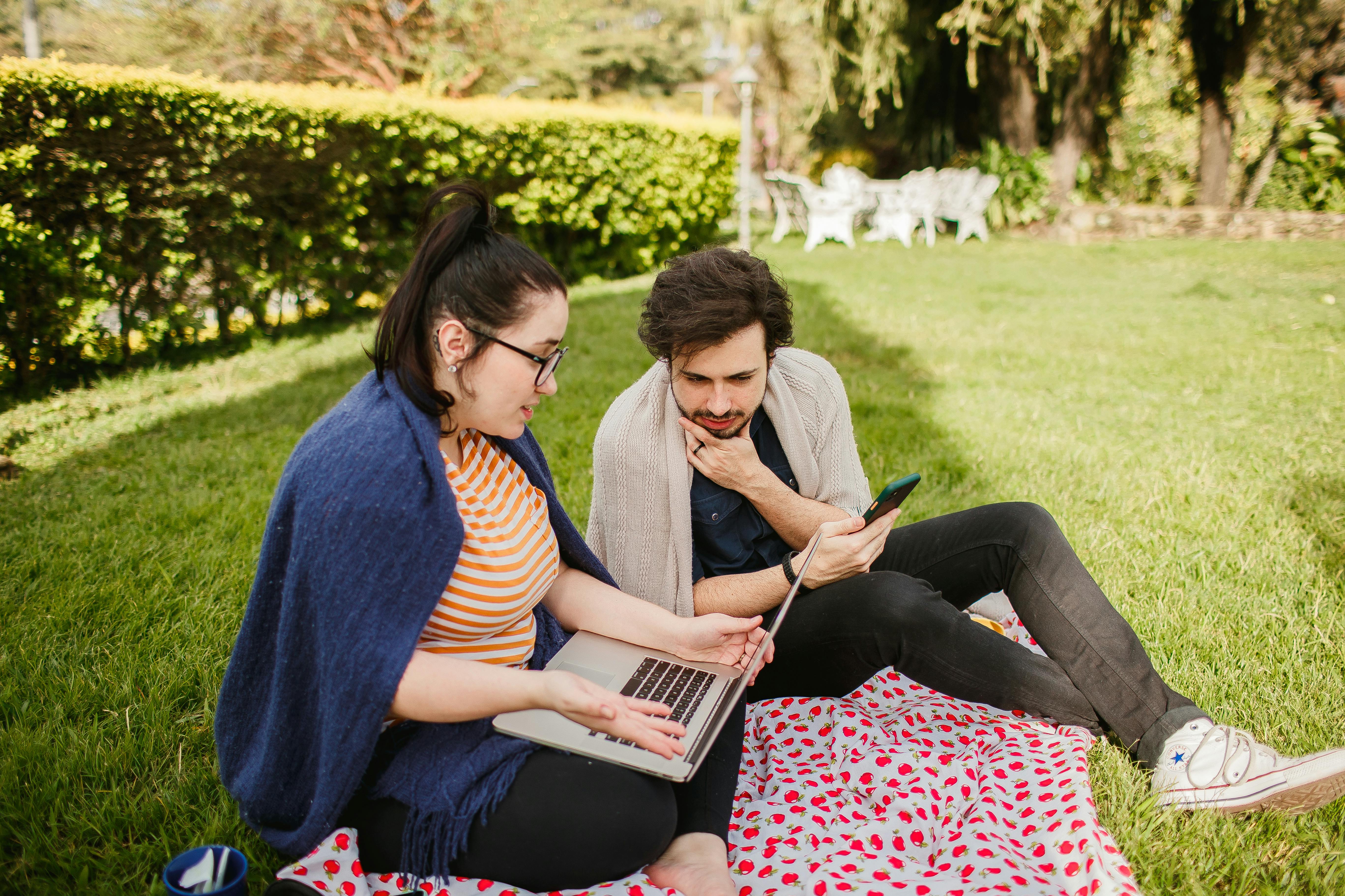 Due giovani seduti sull’erba in un parco, avvolti in coperte e con un laptop appoggiato su una coperta a fantasia, discutono guardando lo schermo come se stessero rivedendo insieme un messaggio o una mail da inviare.