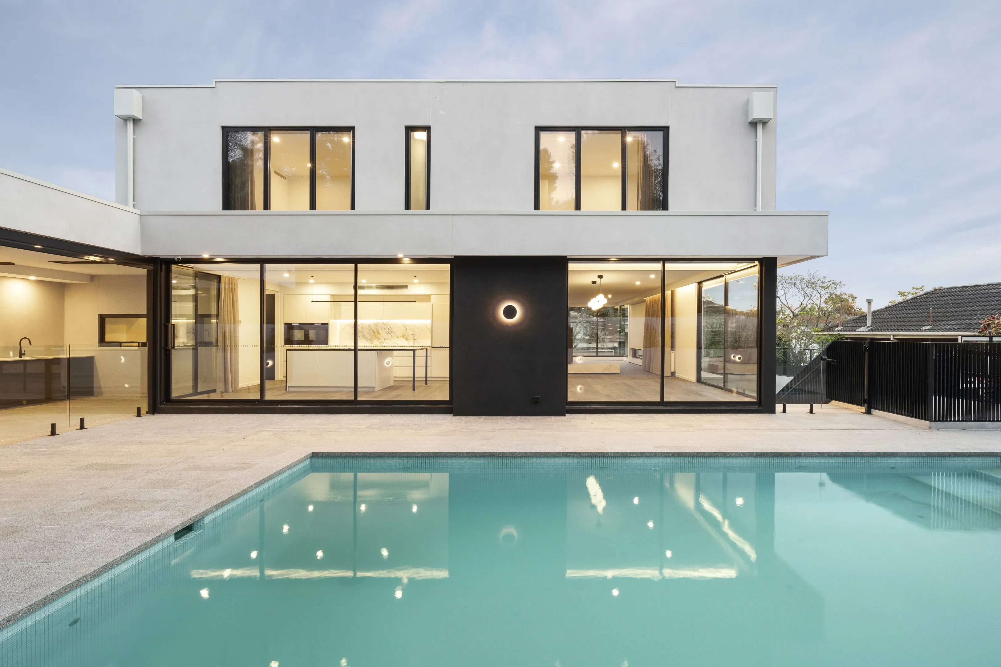 Rear of Ivanhoe East Residence at dusk overlooking a resort-style pool, with a white-rendered upper level, black-framed glazing, a black feature panel and warmly lit interiors