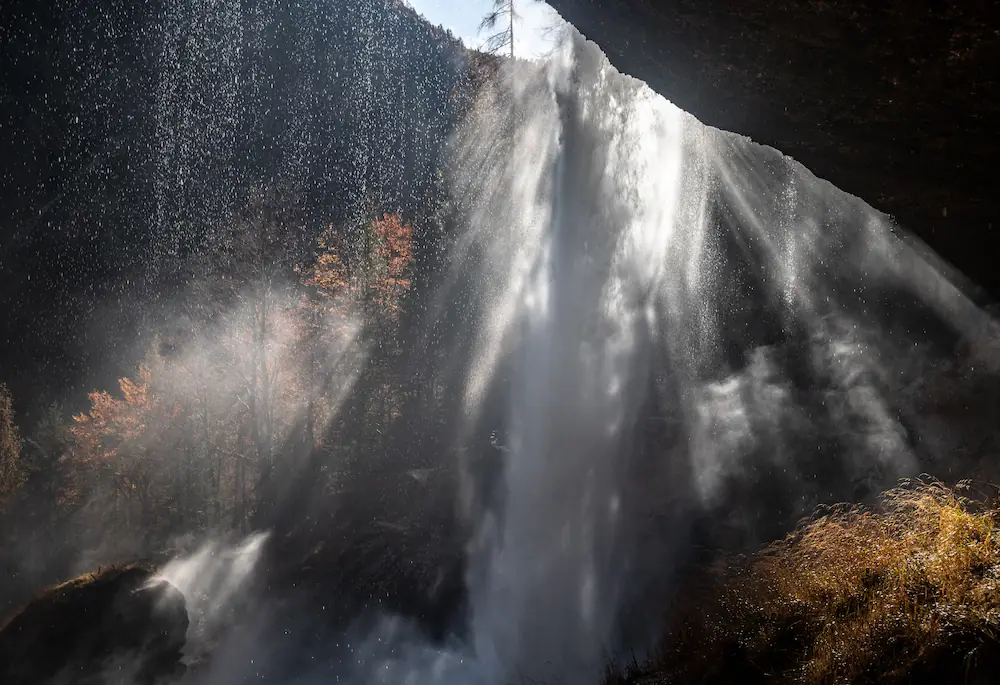 A powerful view from behind Peričnik Waterfall in Slovenia, where the sun illuminates the falling curtain of water, creating bright light rays and sparkling mist against a backdrop of autumn trees and a dark rocky overhang.