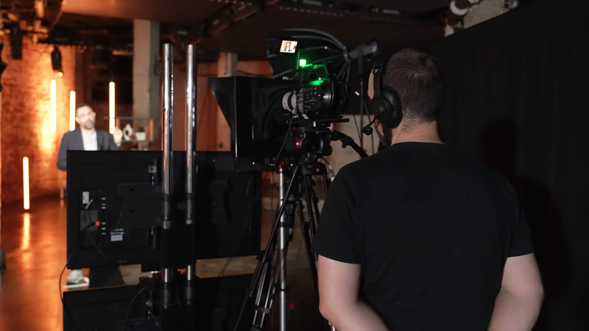 Camera operator filming a keynote speaker in a brick-walled studio environment with warm, cinematic lighting.