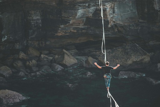 A person wearing a harness walks carefully across a slackline suspended high above rocky coastal waters, with arms stretched out for balance and a cliff face in the background.
