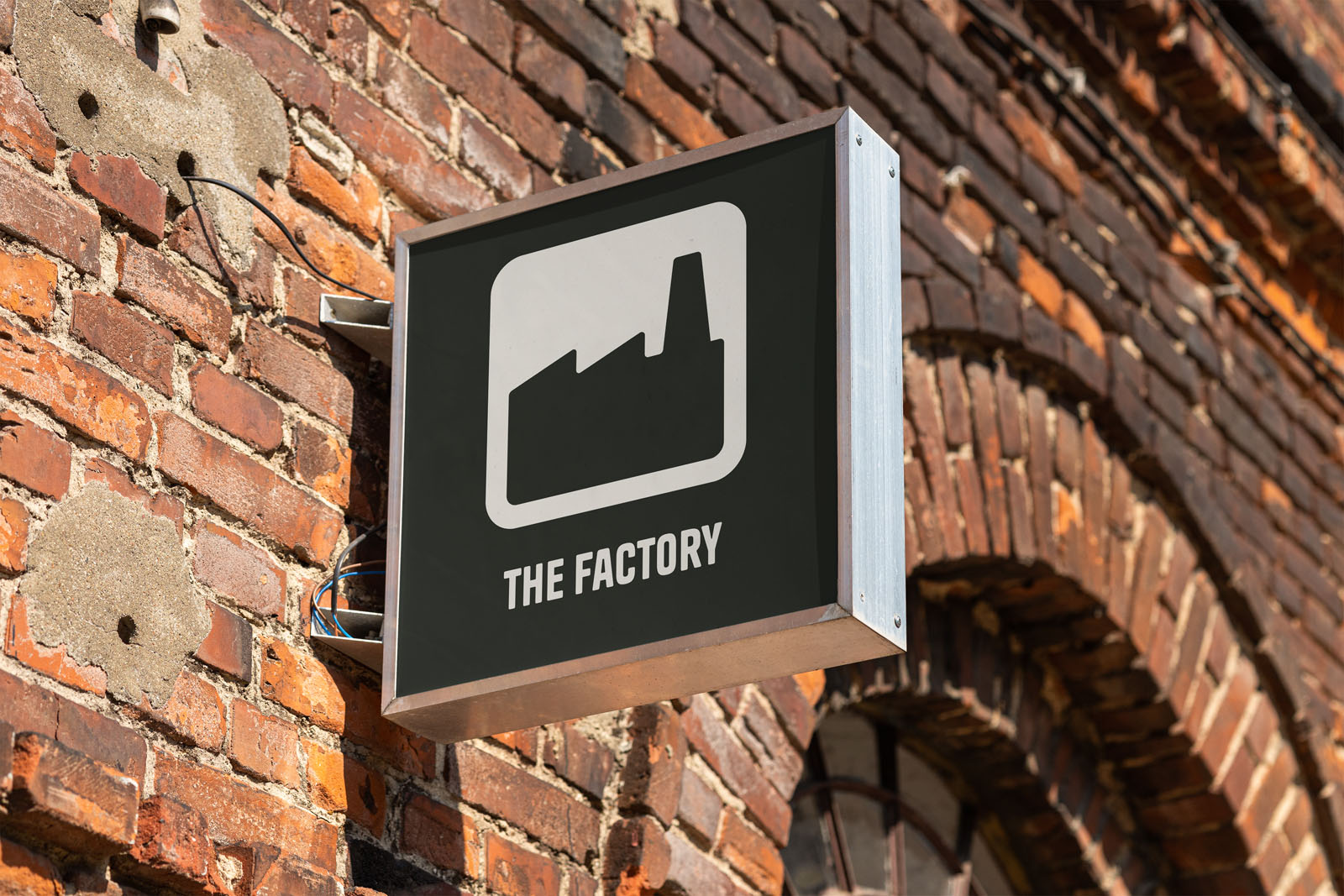 Een verlicht uithangbord aan een karakteristieke rode bakstenen muur. Het bord toont een wit icoon van een fabrieksgebouw met de tekst "THE FACTORY".