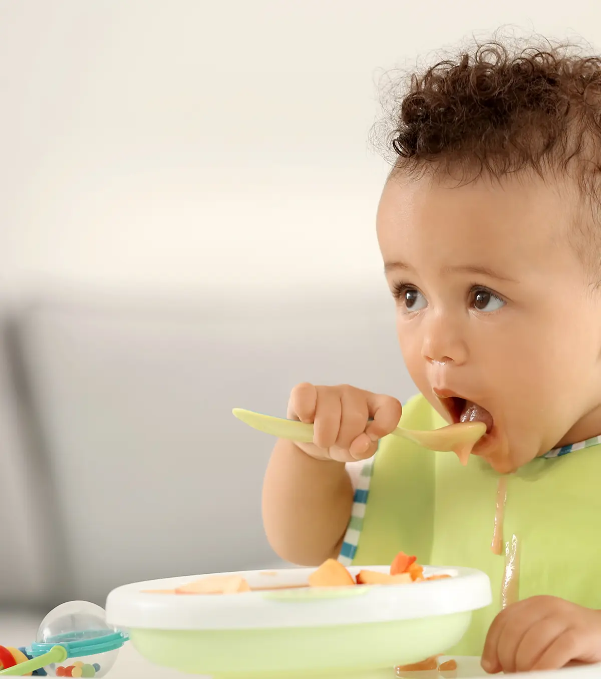 Baby wearing a green bib eating soft food with a spoon at a high chair.