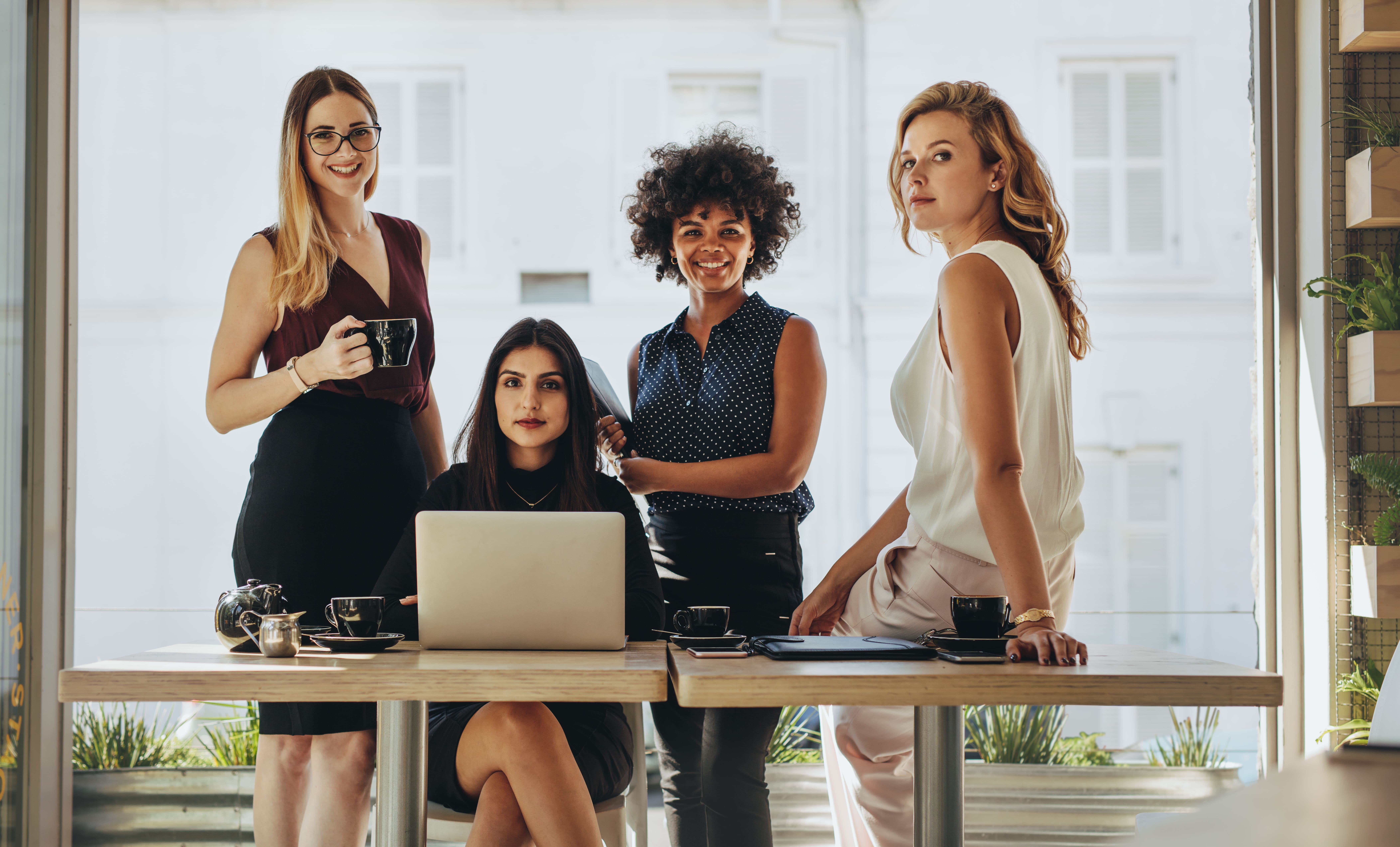 Imagem de quatro mulheres em um ambiente de trabalho, representando diversidade e profissionalismo. Elas estão reunidas ao redor de uma mesa, onde há um laptop, xícaras de café e acessórios de escritório. Uma delas está sentada, enquanto as outras três permanecem em pé, transmitindo uma atmosfera de colaboração e confiança. A iluminação suave e o fundo com janelas amplas realçam a harmonia do ambiente.