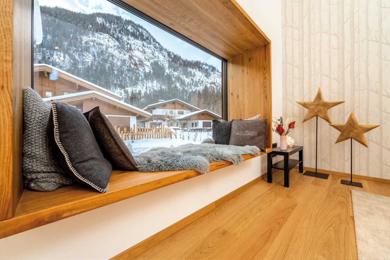 Fensterbank Sitznische mit Kissen und Schaffell, Blick auf schneebedeckte Berge und Häuser, Holzfußboden und Baum-Tapete.