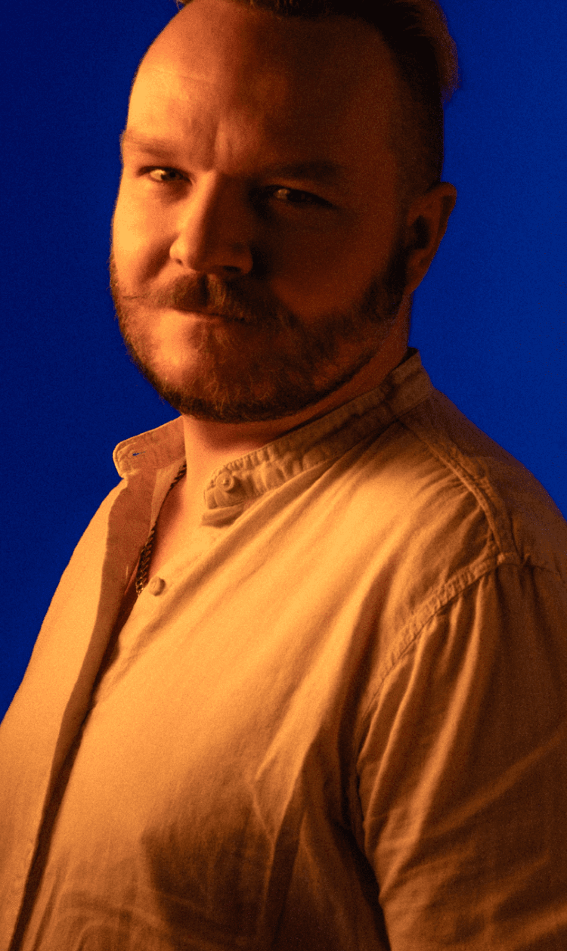 Zbliżenie na twarz muzyka z formacji Electric Shapes, z charakterystycznymi wąsami i w teksturowanej koszuli, skąpanego w świetle o barwie złotej godziny na minimalistycznym niebieskim tle.